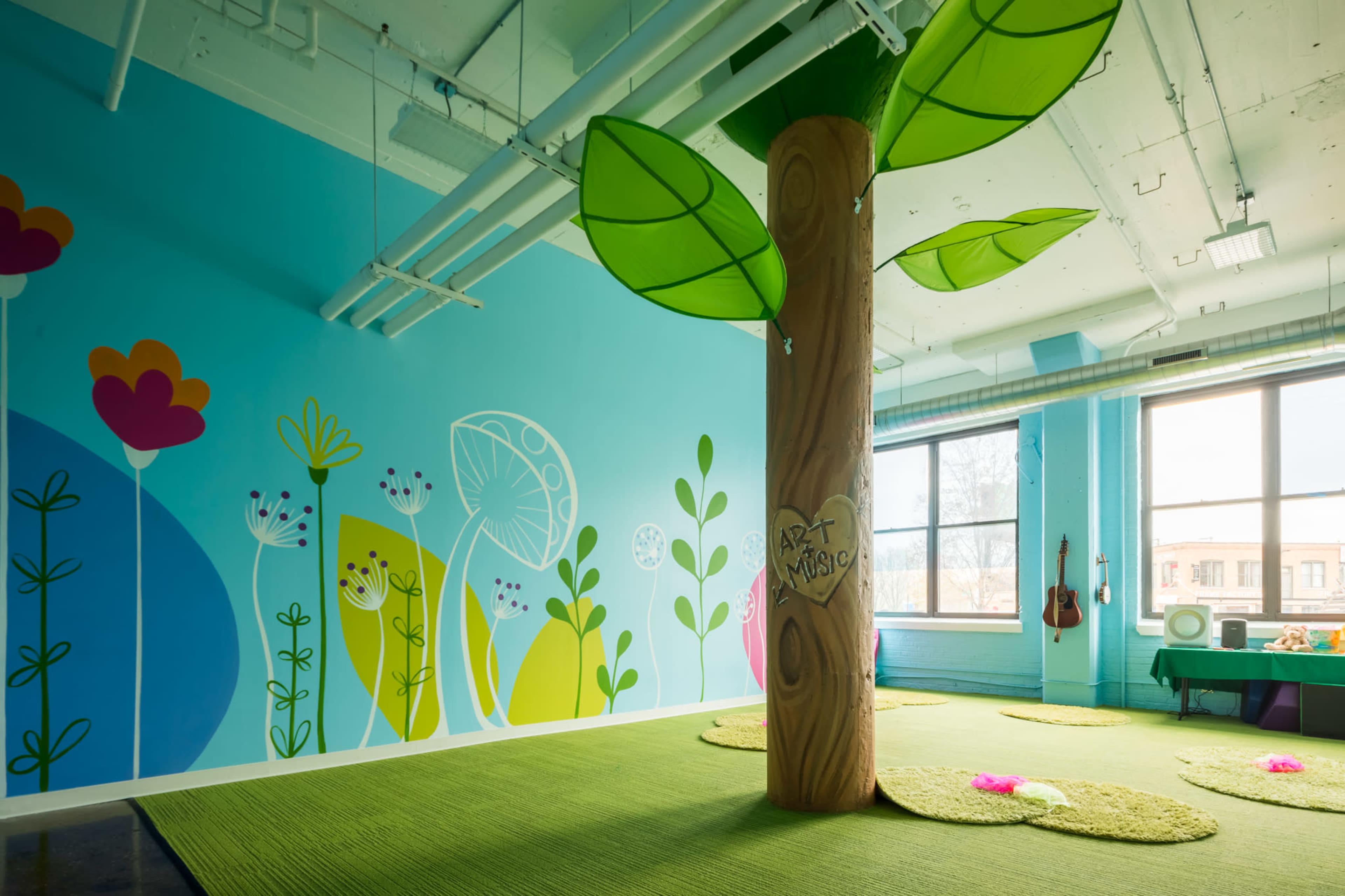 A brightly colored playroom featuring a large tree structure with green leaves and a mural of flowers and plants on the walls.