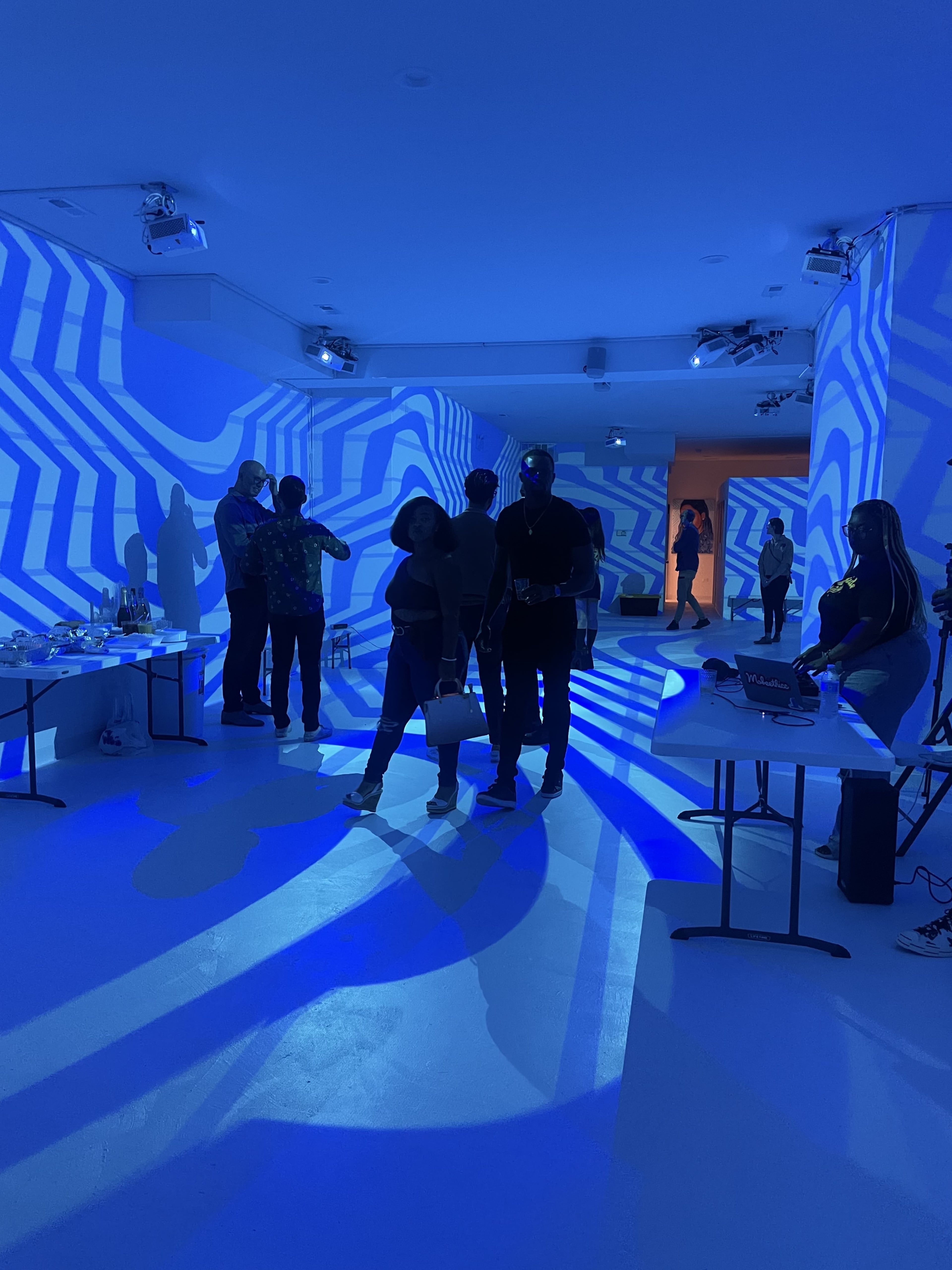 A gallery space with people engaged in conversation, while colorful geometric projections cover the walls and floor.