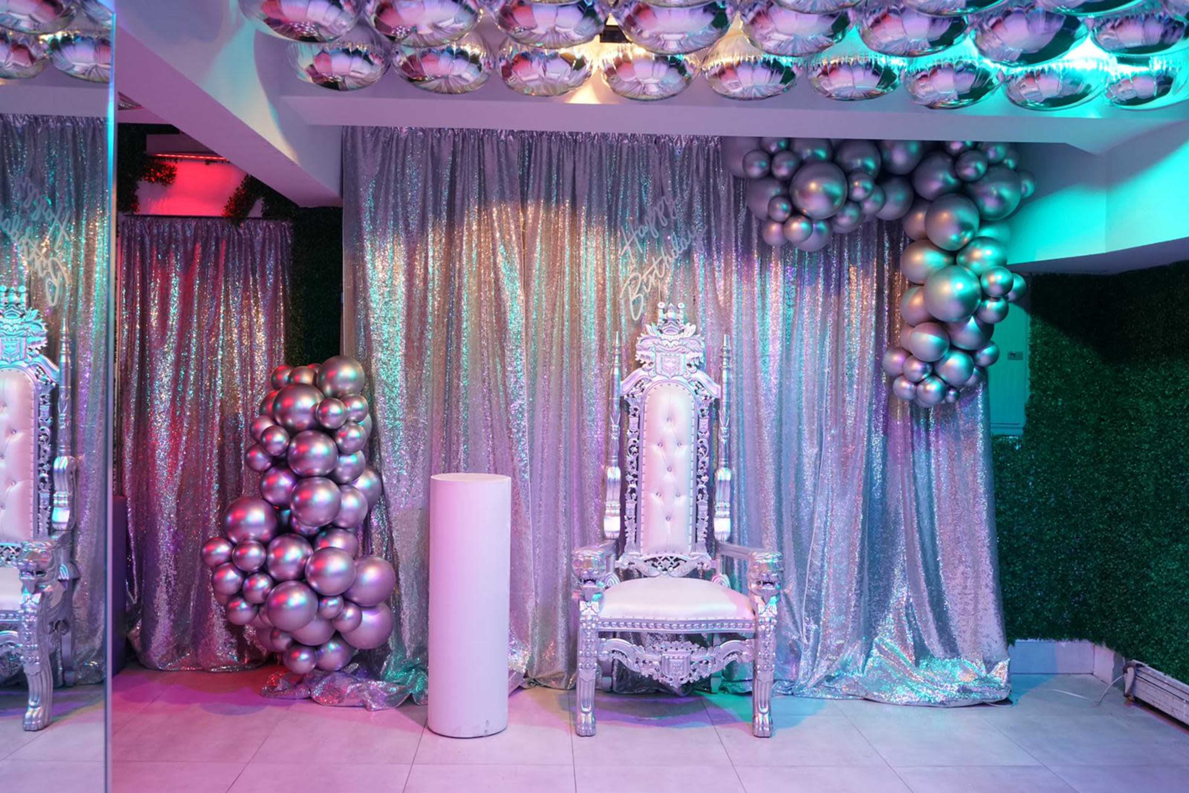 A decorative indoor space with a glittering silver backdrop, a large ornate throne chair, and clusters of metallic balloons.