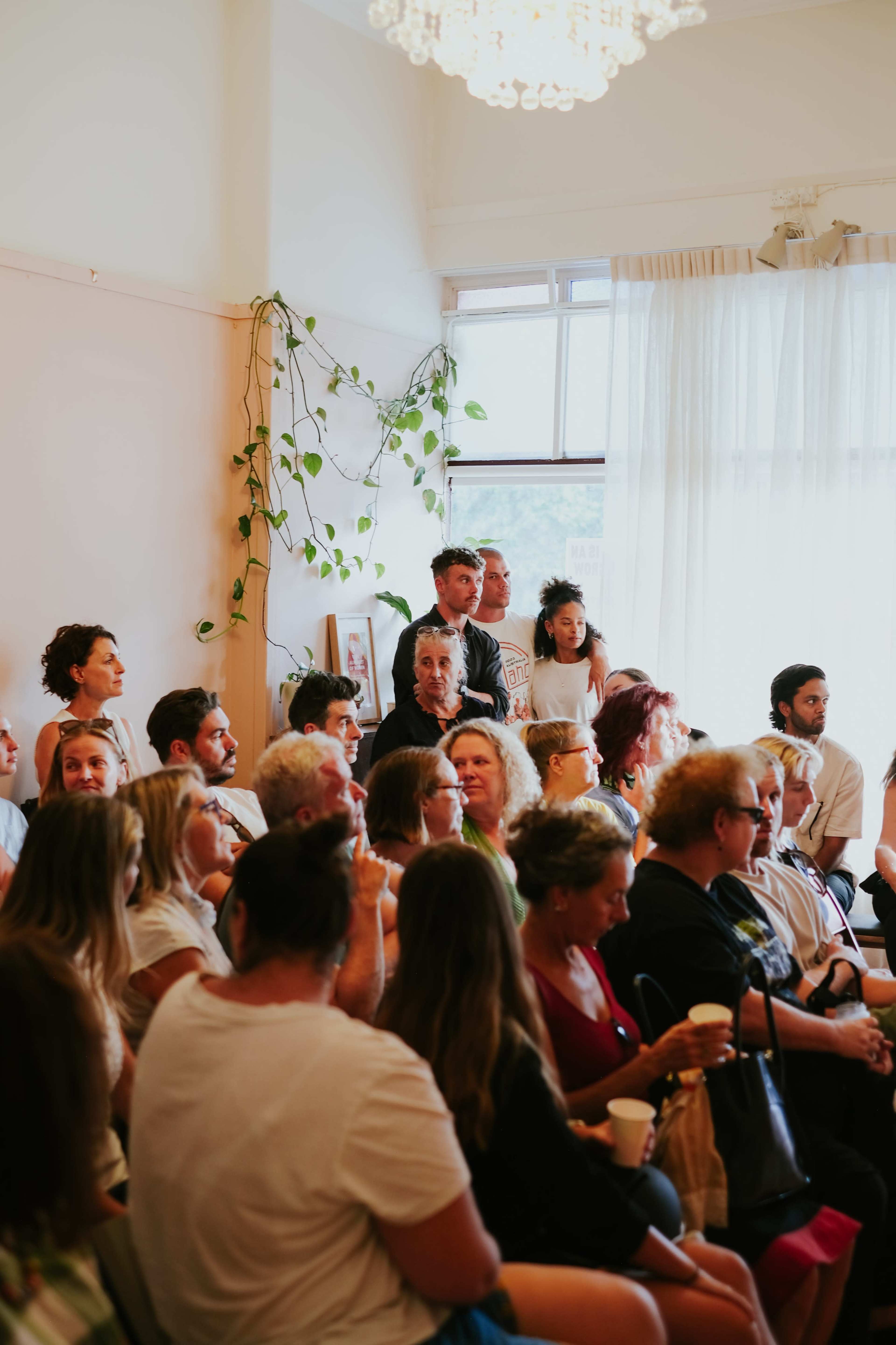 A diverse group of people is seated and standing in a well-lit room, with plants adorning the walls and sunlight streaming through the windows.