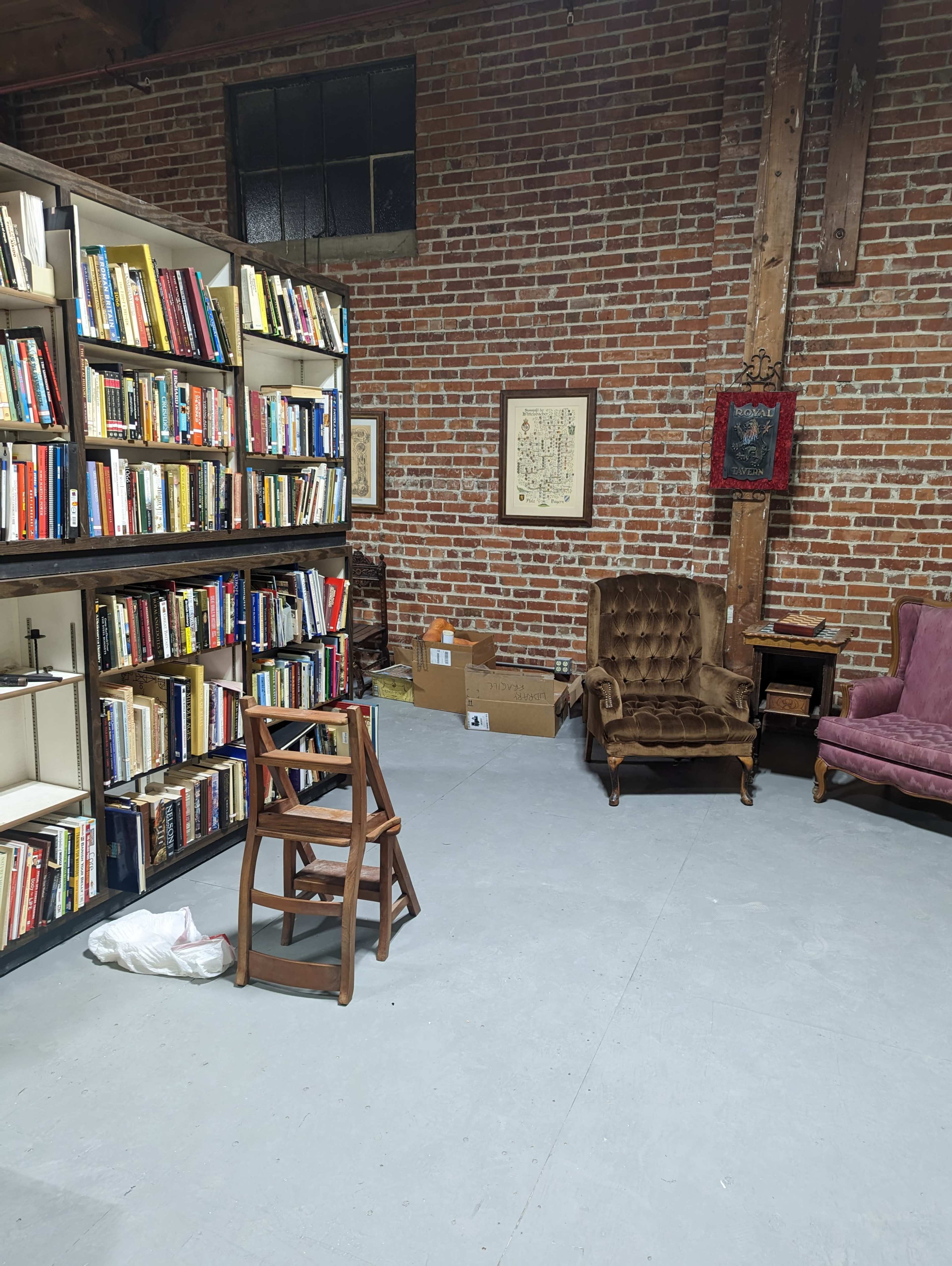 A room with brick walls, featuring a bookshelf filled with books, two armchairs, a stack of boxes, and a wooden chair in the center.