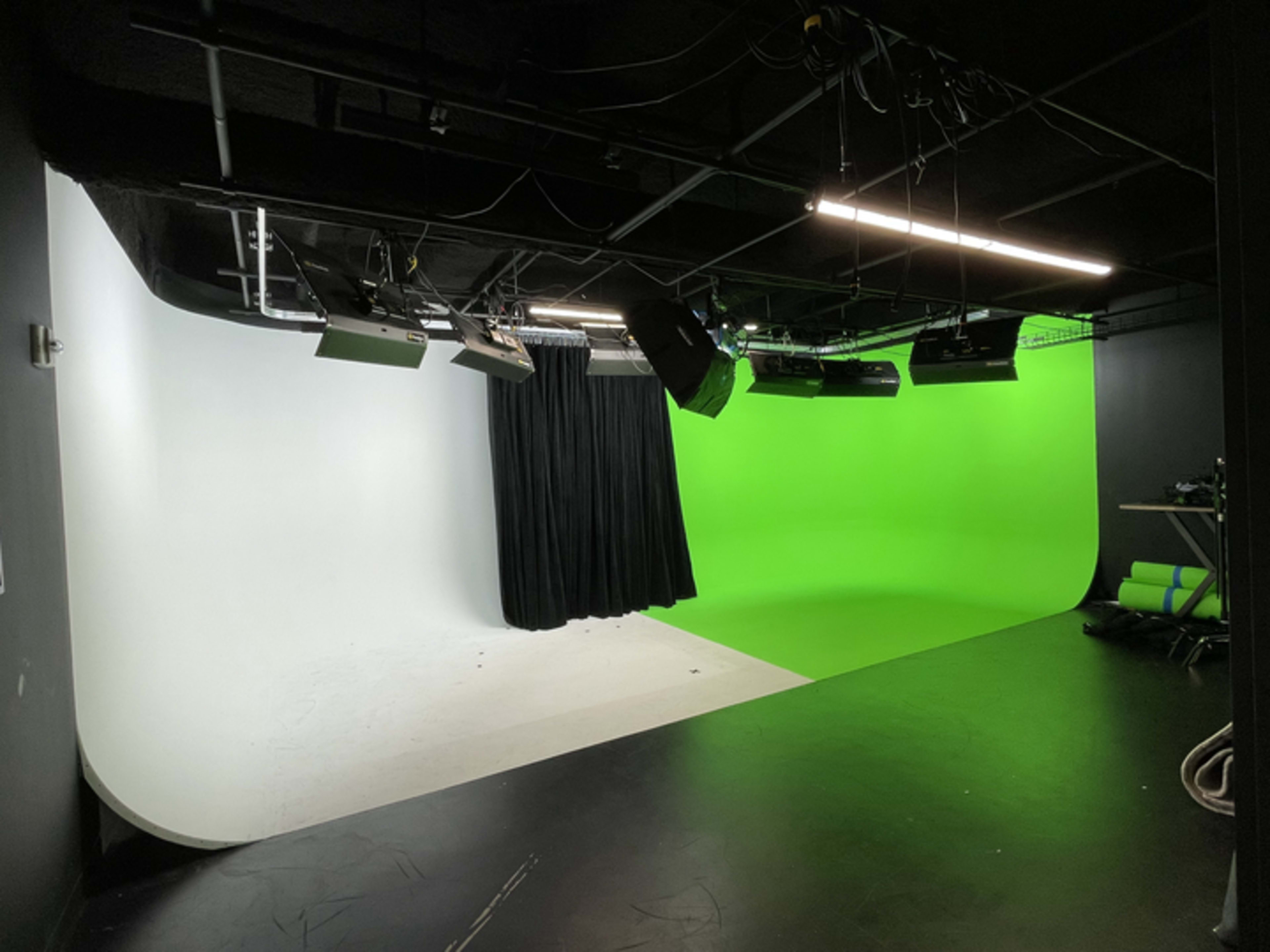 A studio with a split backdrop, featuring a white side and a green screen side, equipped with various lighting equipment and a black curtain.