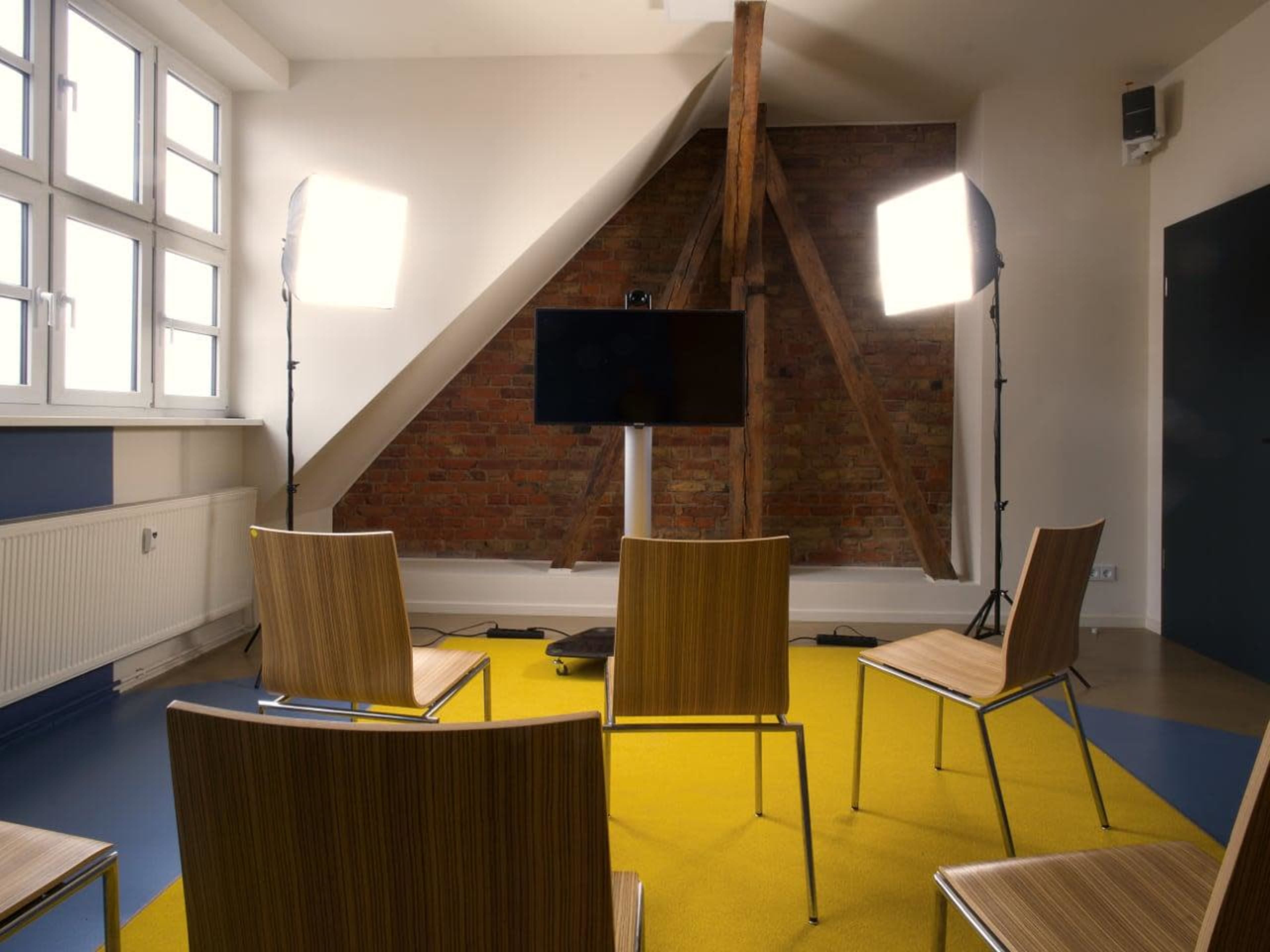 A meeting room is set up with four wooden chairs facing a television screen, illuminated by two bright lights, and featuring a yellow carpet with exposed brick walls.