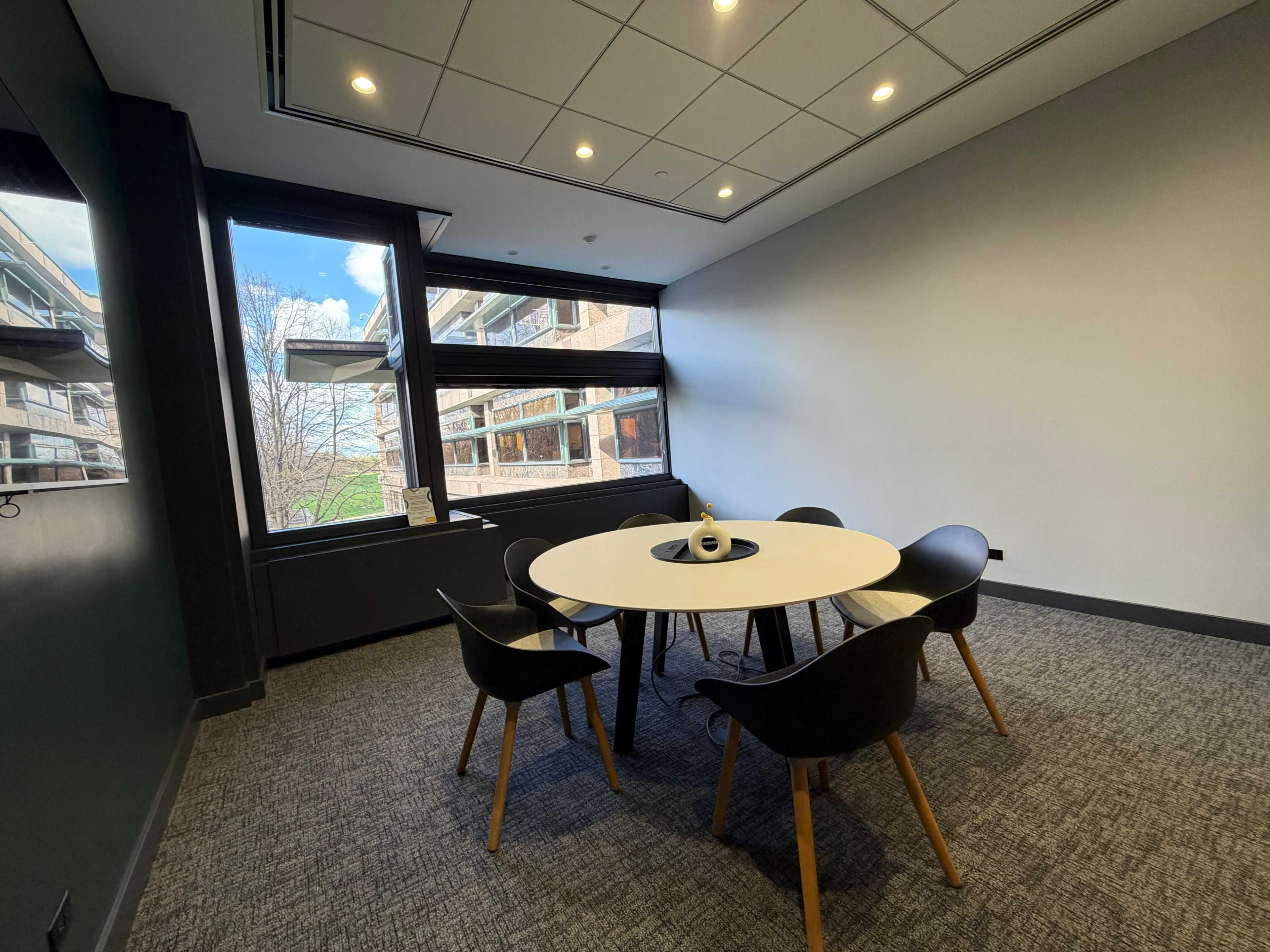 A small meeting room features a round table surrounded by five black chairs, with large windows offering a view of an exterior building and trees.