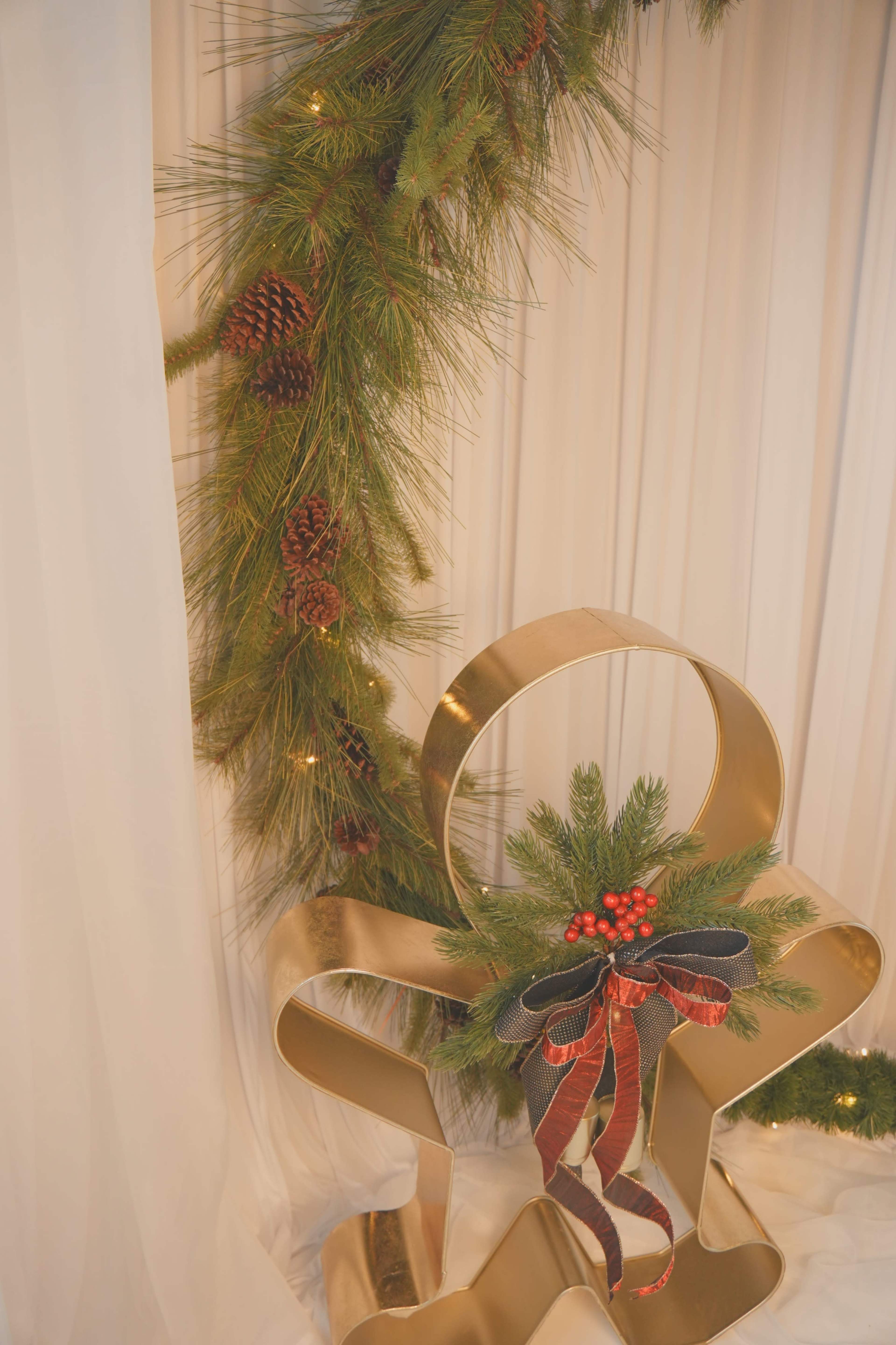 A gold, gingerbread-shaped chair is decorated with evergreen branches, red berries, and a black ribbon, positioned near a pine garland.