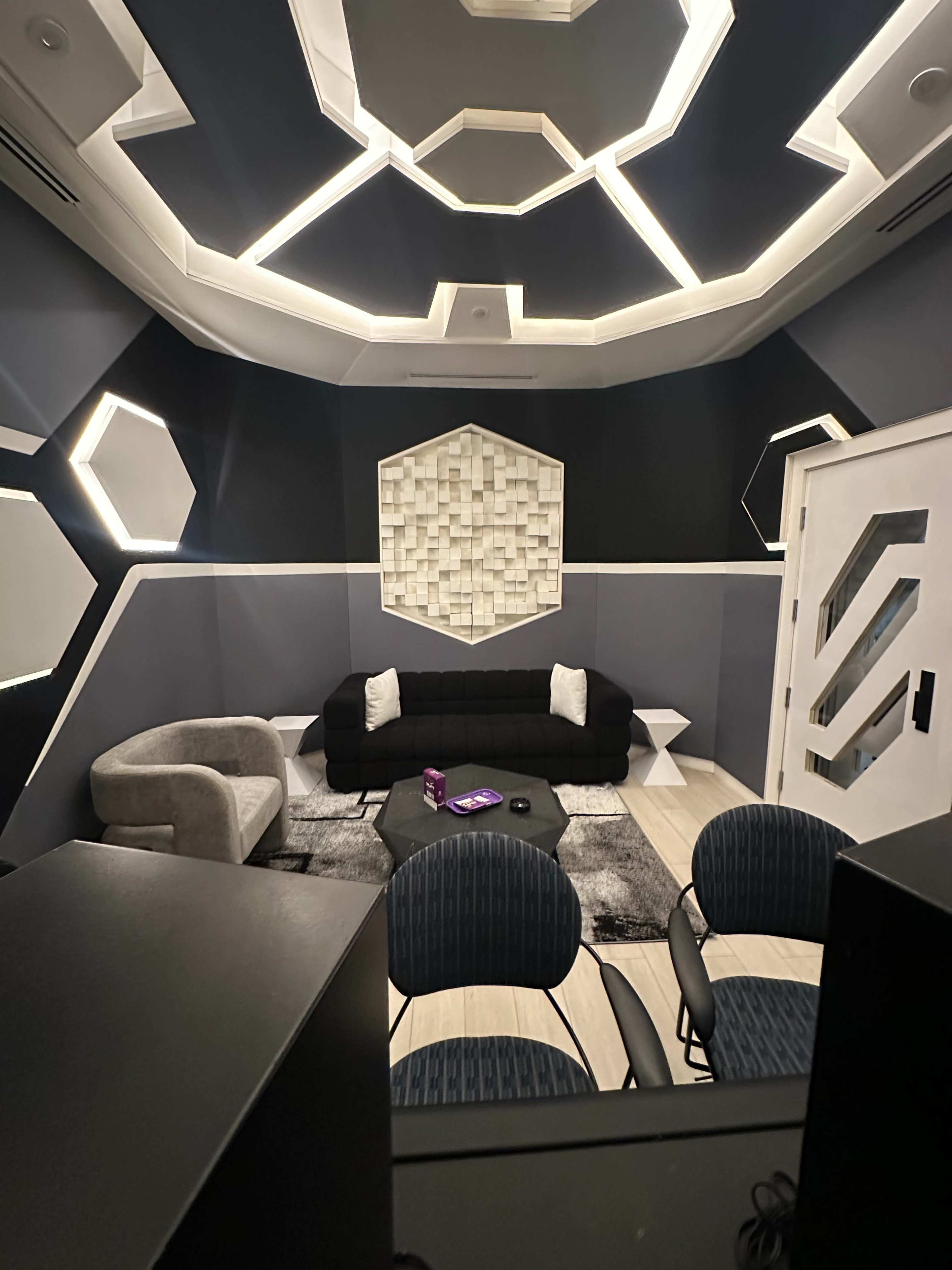 The image shows a modern, geometric interior with a black couch, light-colored accent furniture, and a ceiling featuring a patterned design.