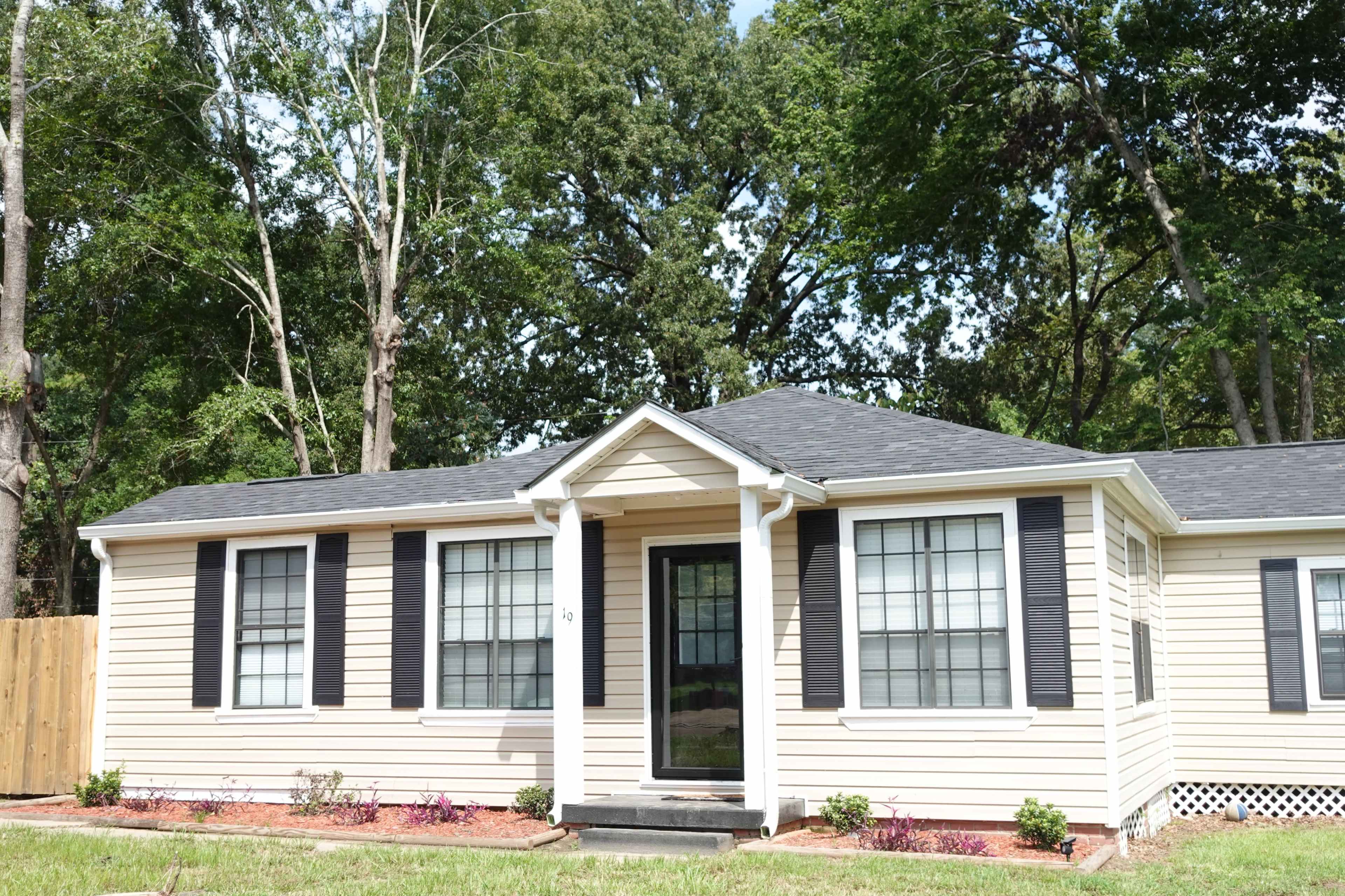 A single-story, beige house with black shutters, a small front porch, and a well-maintained lawn surrounded by trees.