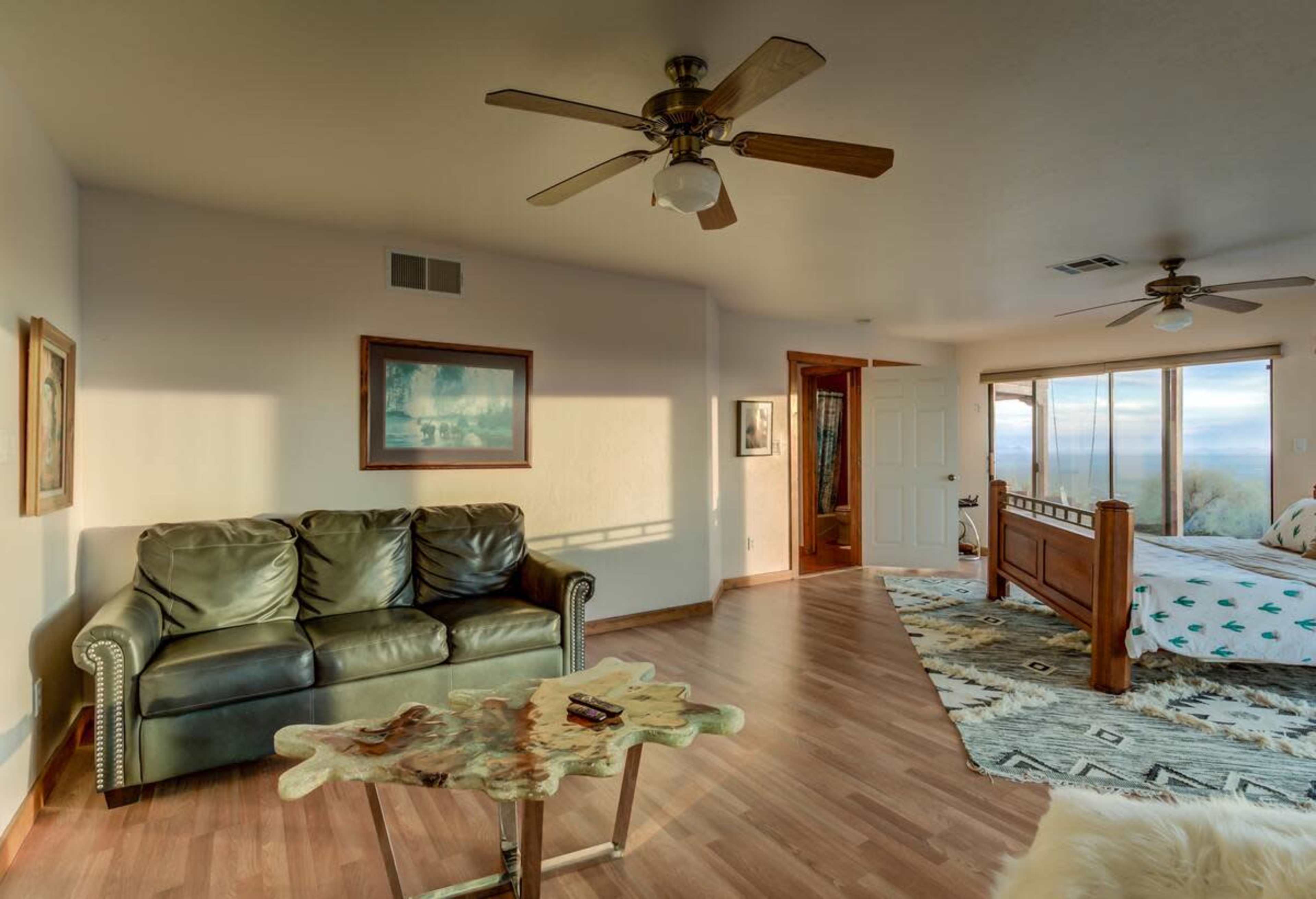 The image shows a spacious bedroom with a large window, a bed, a leather sofa, and a wooden coffee table on a patterned rug.