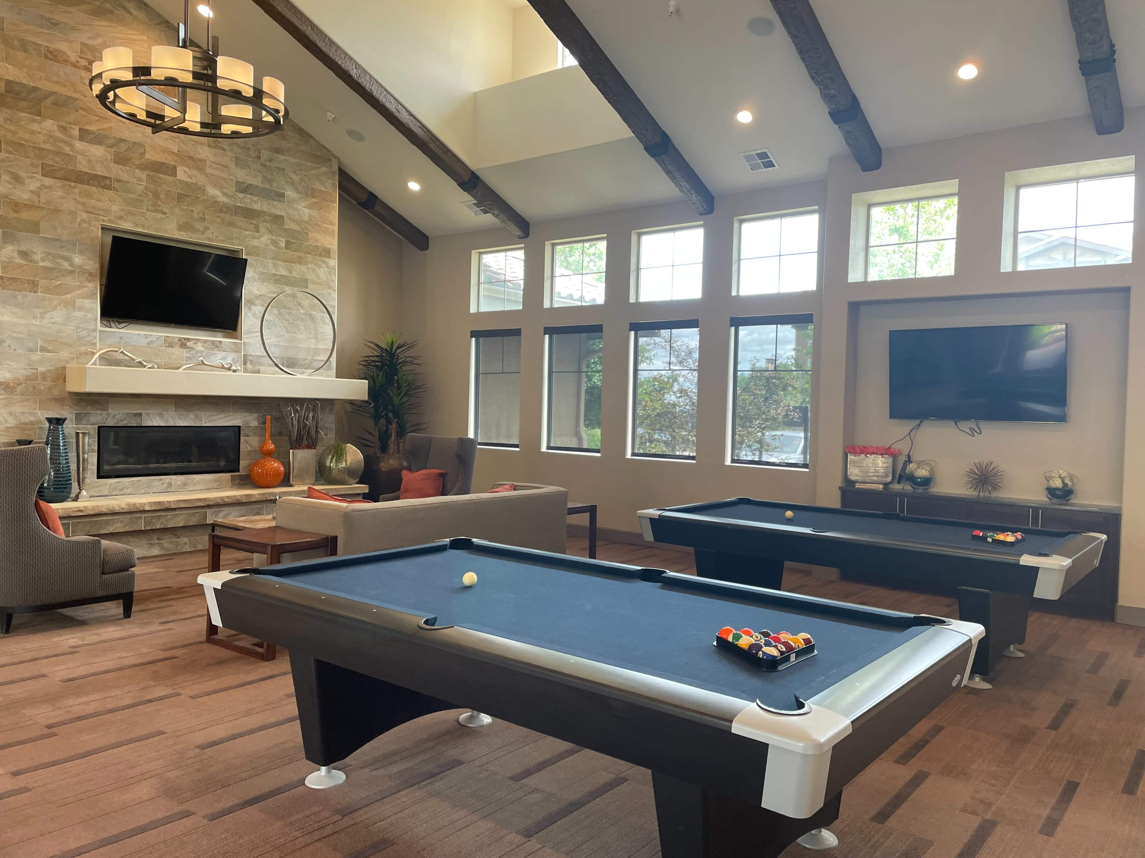 The image shows a spacious lounge area featuring two pool tables, a stone fireplace, and large windows letting in natural light.