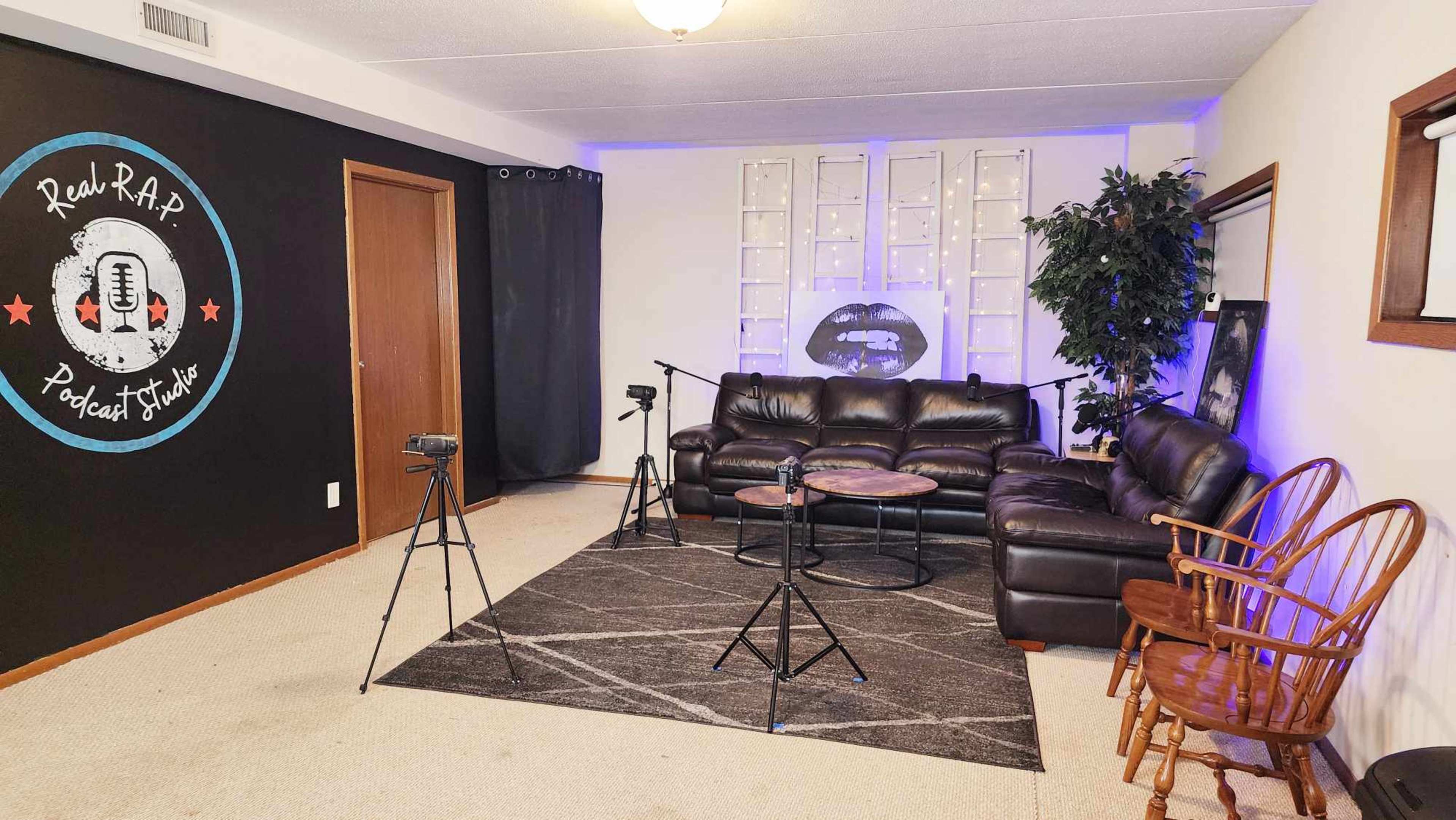 The image shows a podcast studio featuring a seating area with a dark couch, a round table, and two microphones on stands, against a backdrop of blue lights and decorative elements.