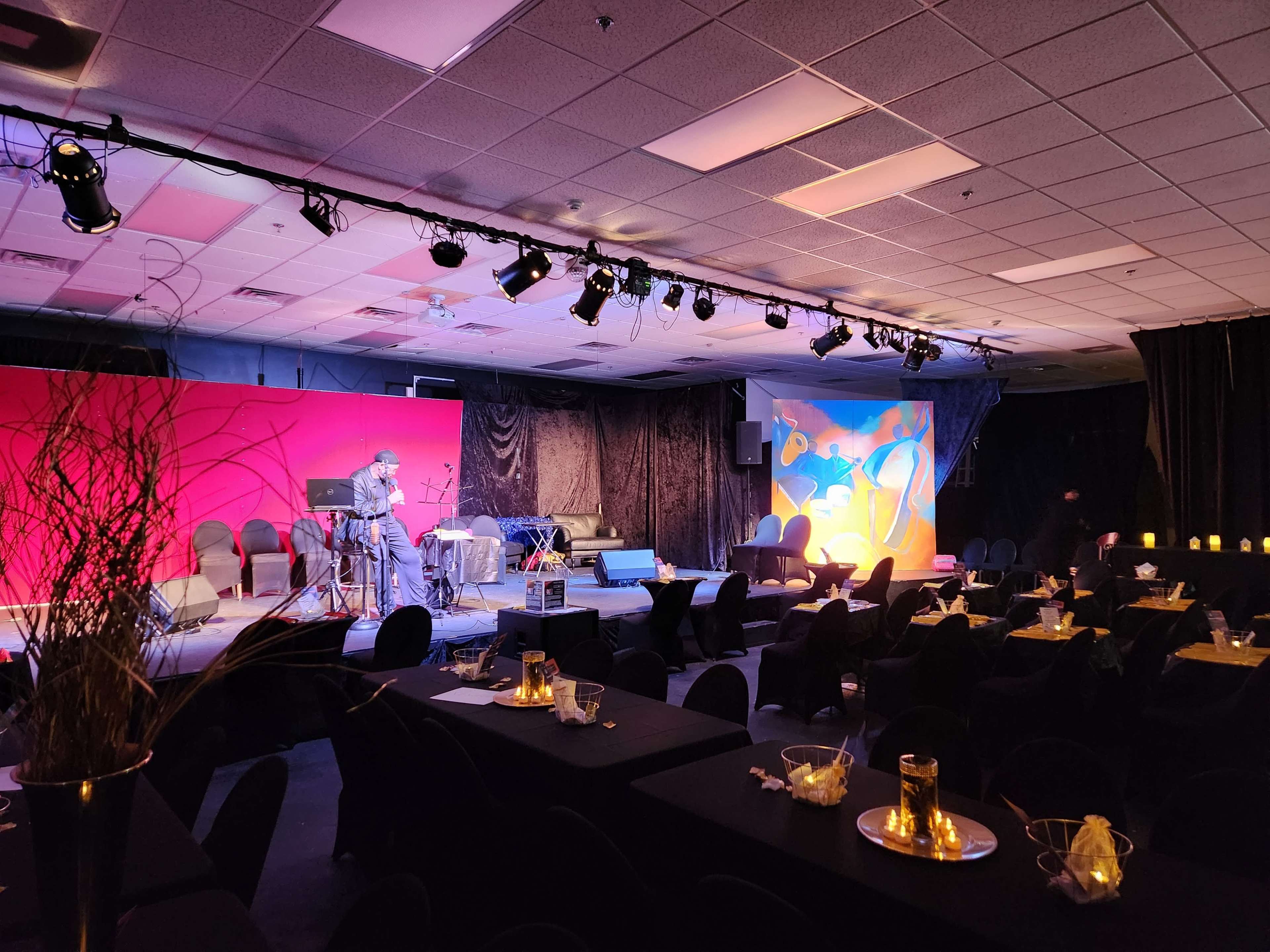 A stage with a musician performing is set up at the front of a dimly lit room filled with tables and chairs.