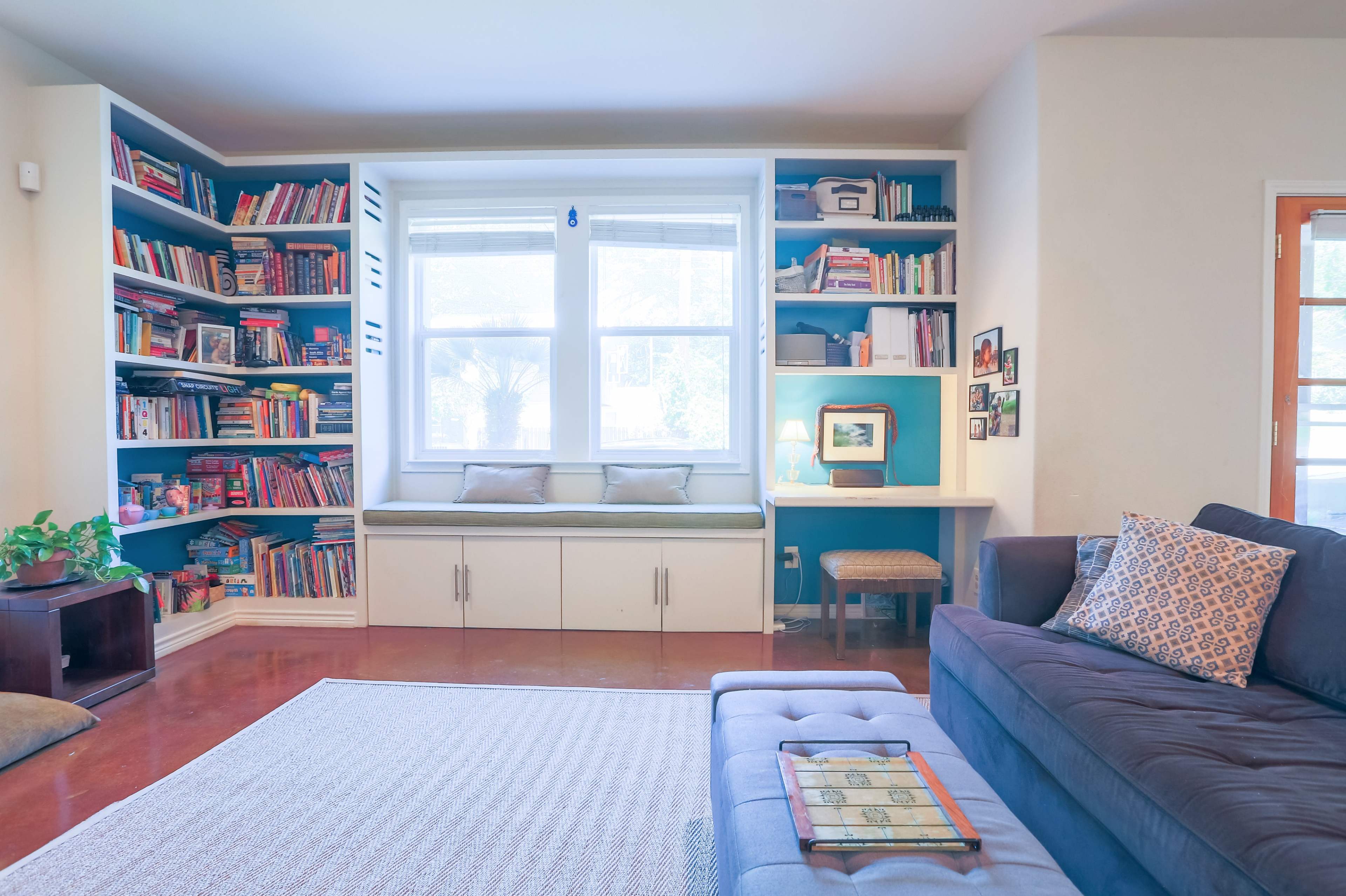 A cozy living room features a large bookshelf filled with colorful books, a window seat with cushions, and a small desk with a picture frame.