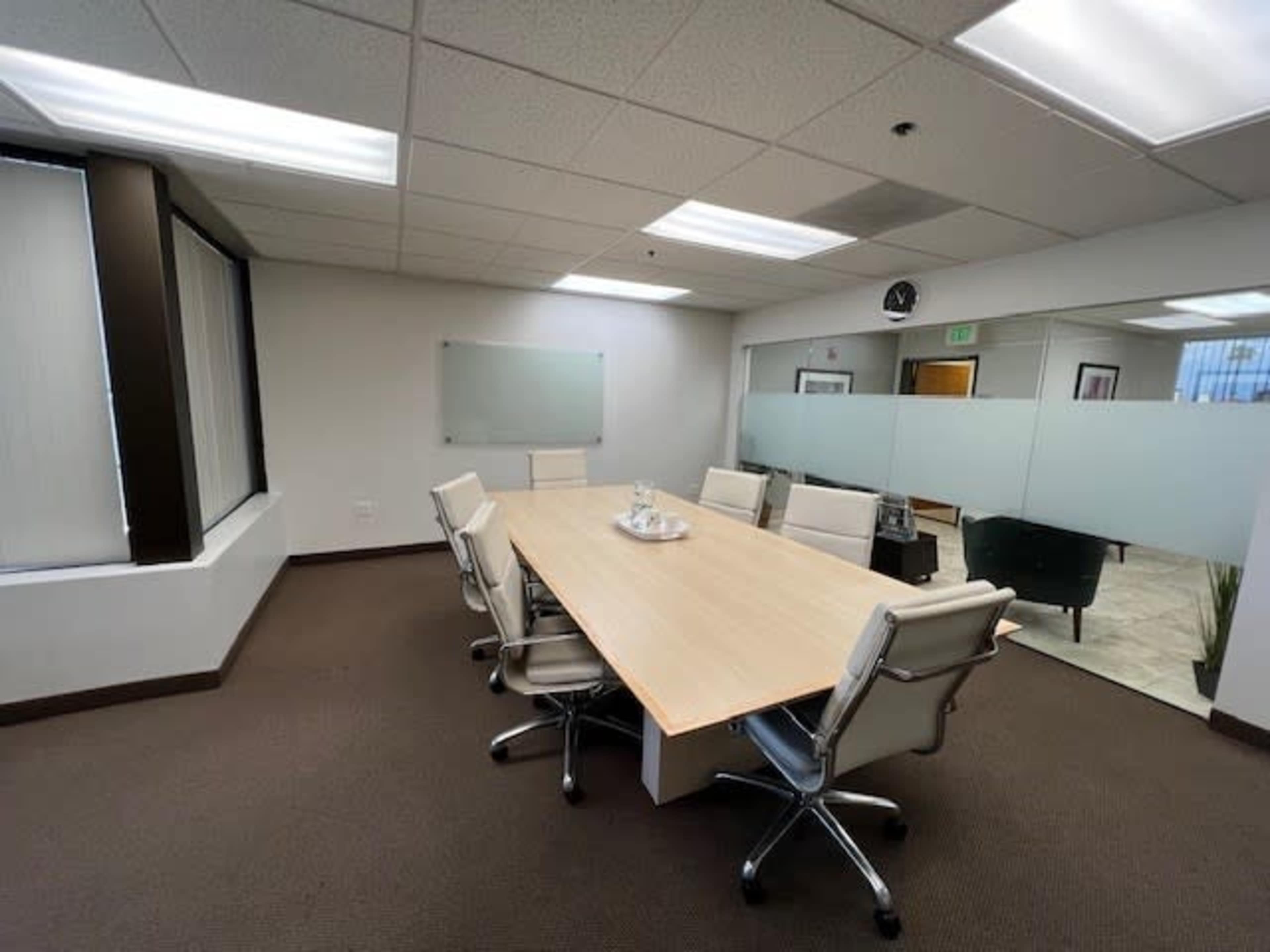 A conference room features a long wooden table surrounded by six white chairs, with a glass whiteboard and large windows on two sides.