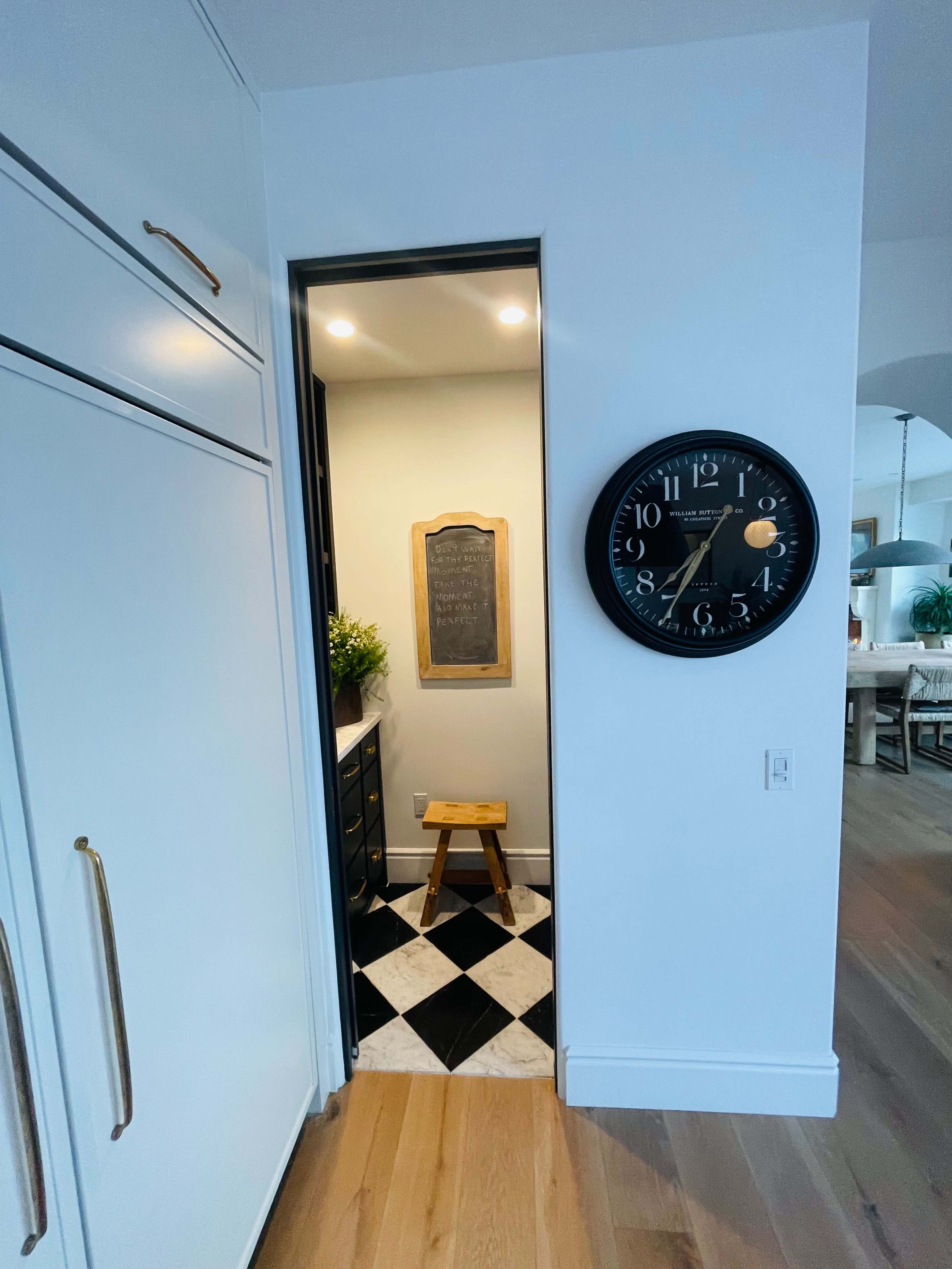 A narrow hallway with a black and white checkered floor leads to a small room that features a chalkboard and a wooden stool, alongside a large wall clock.