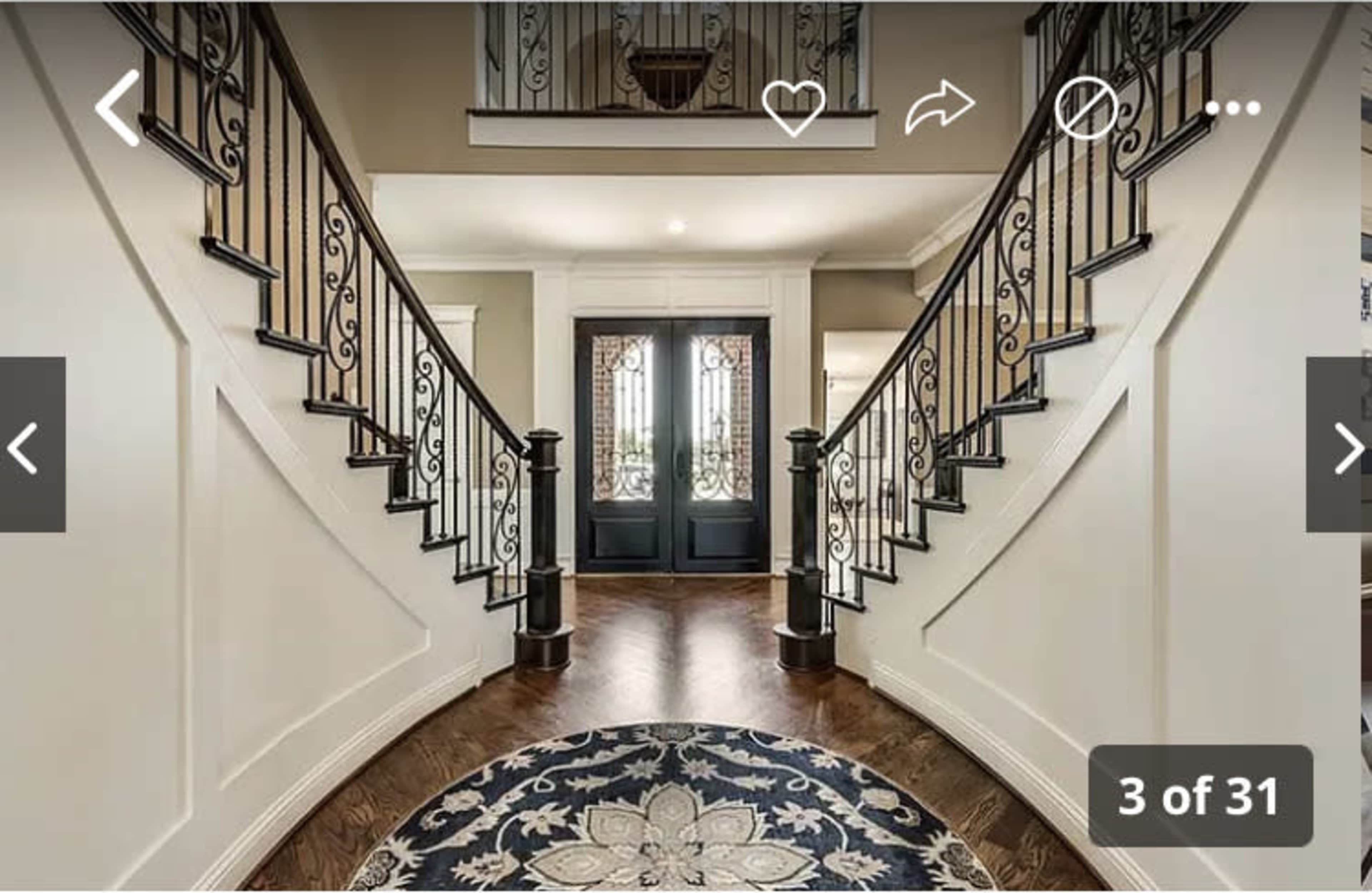 The image shows a spacious foyer with a grand staircase on either side, leading to double doors with decorative glass inserts.