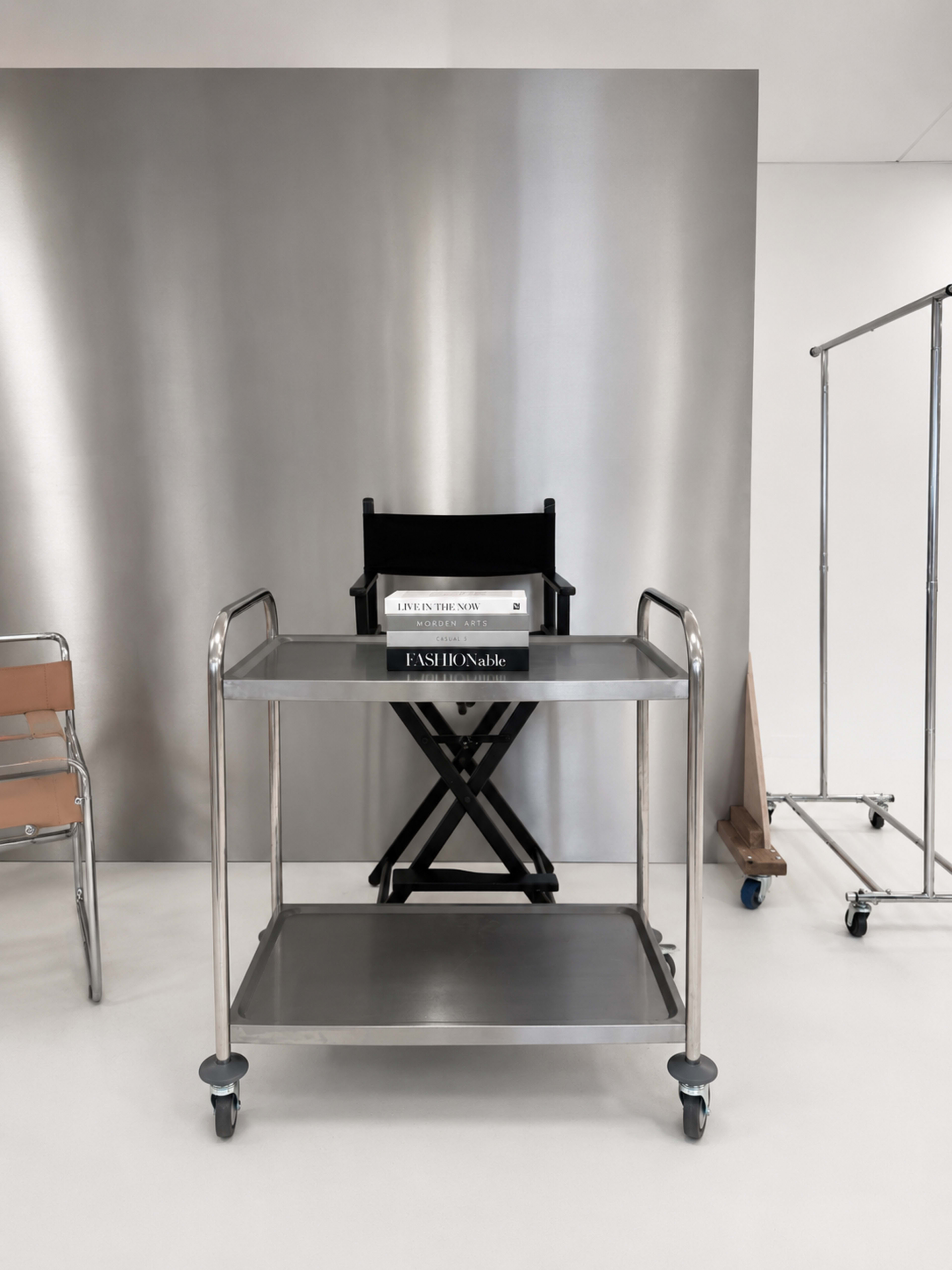 A metallic serving cart is positioned in front of a sleek, silver backdrop, with a director's chair and a stack of books on top.
