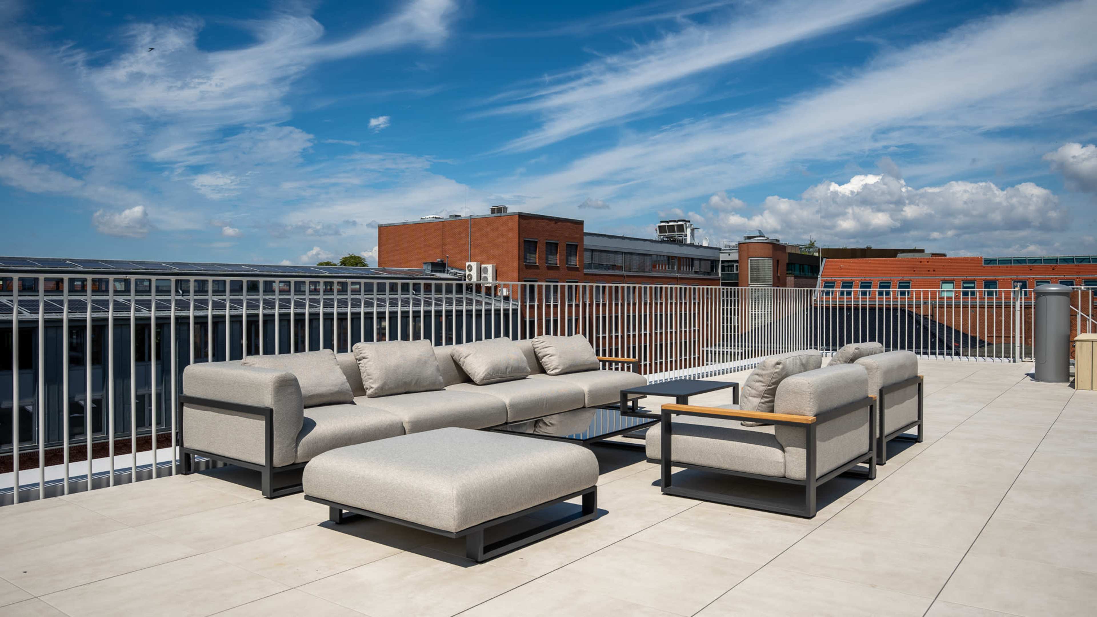 The image shows a rooftop lounge area featuring modern, light-colored sectional sofas and an ottoman on a tiled surface, with a clear blue sky overhead.