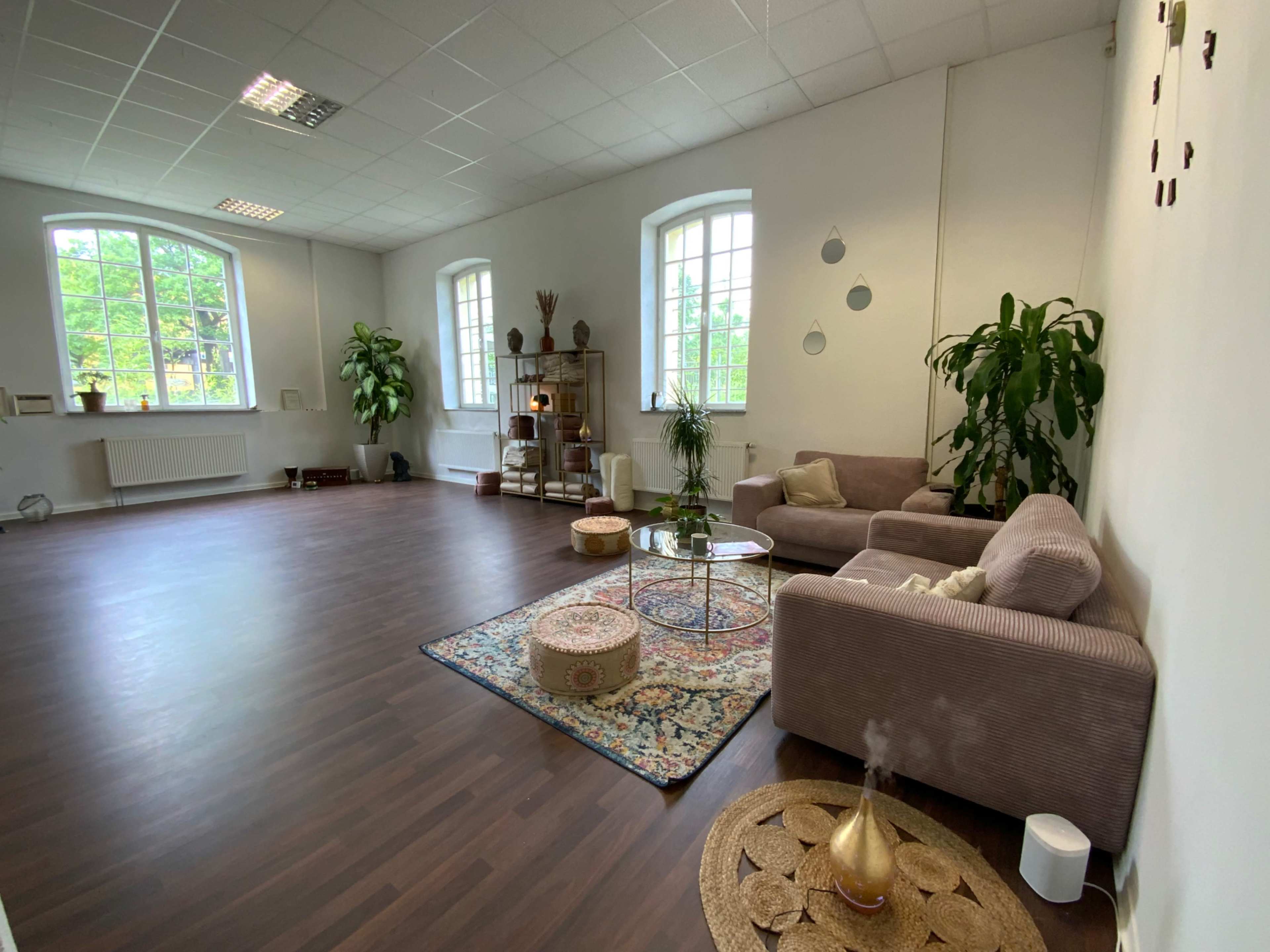 The image shows a spacious room with wooden flooring, large windows, plants, a sofa, a coffee table, and decorative elements including a rug and poufs.