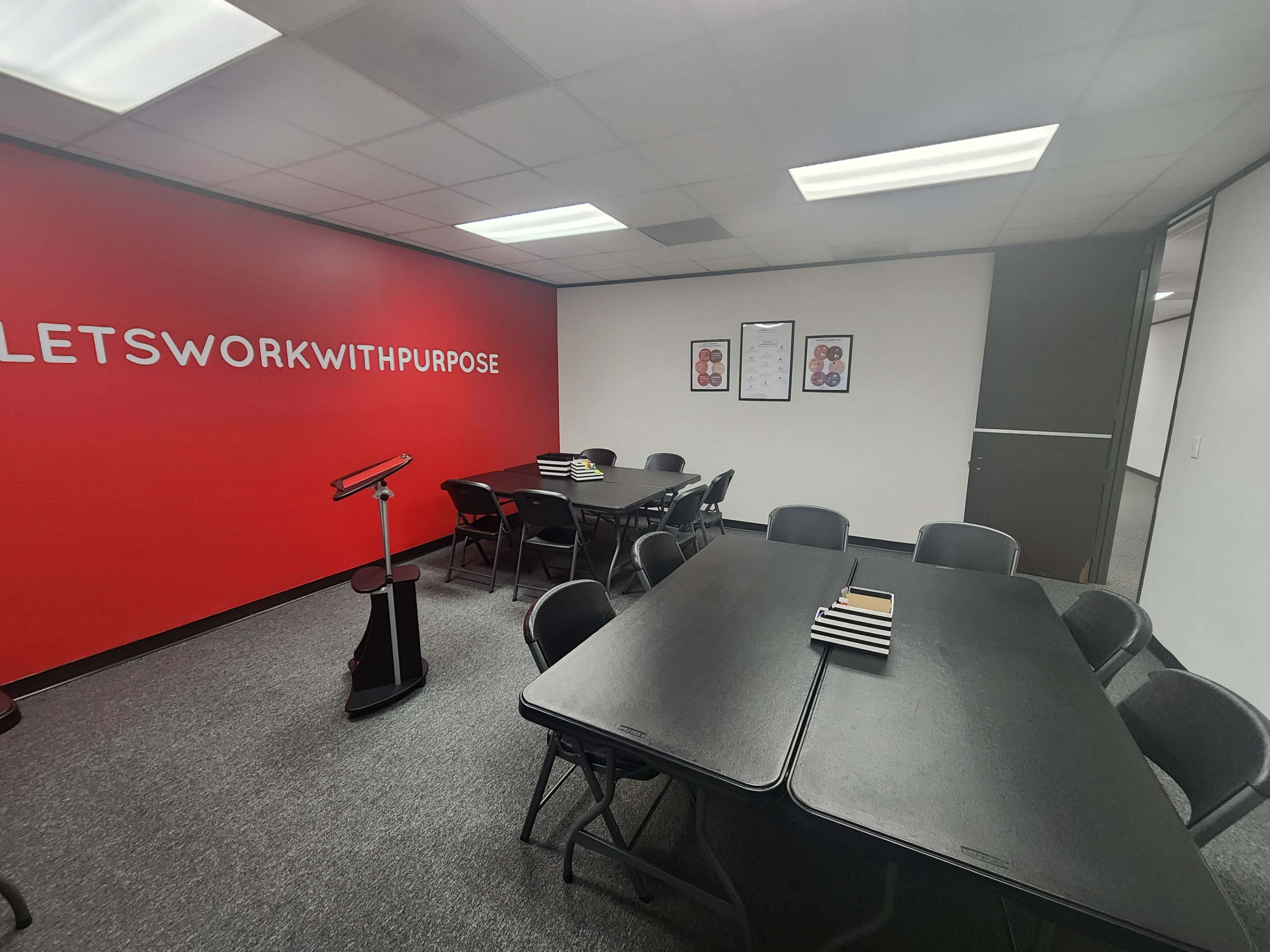 The image shows a conference room with several black folding tables and chairs, a podium, and a bright red wall featuring the phrase "LETS WORK WITH PURPOSE."