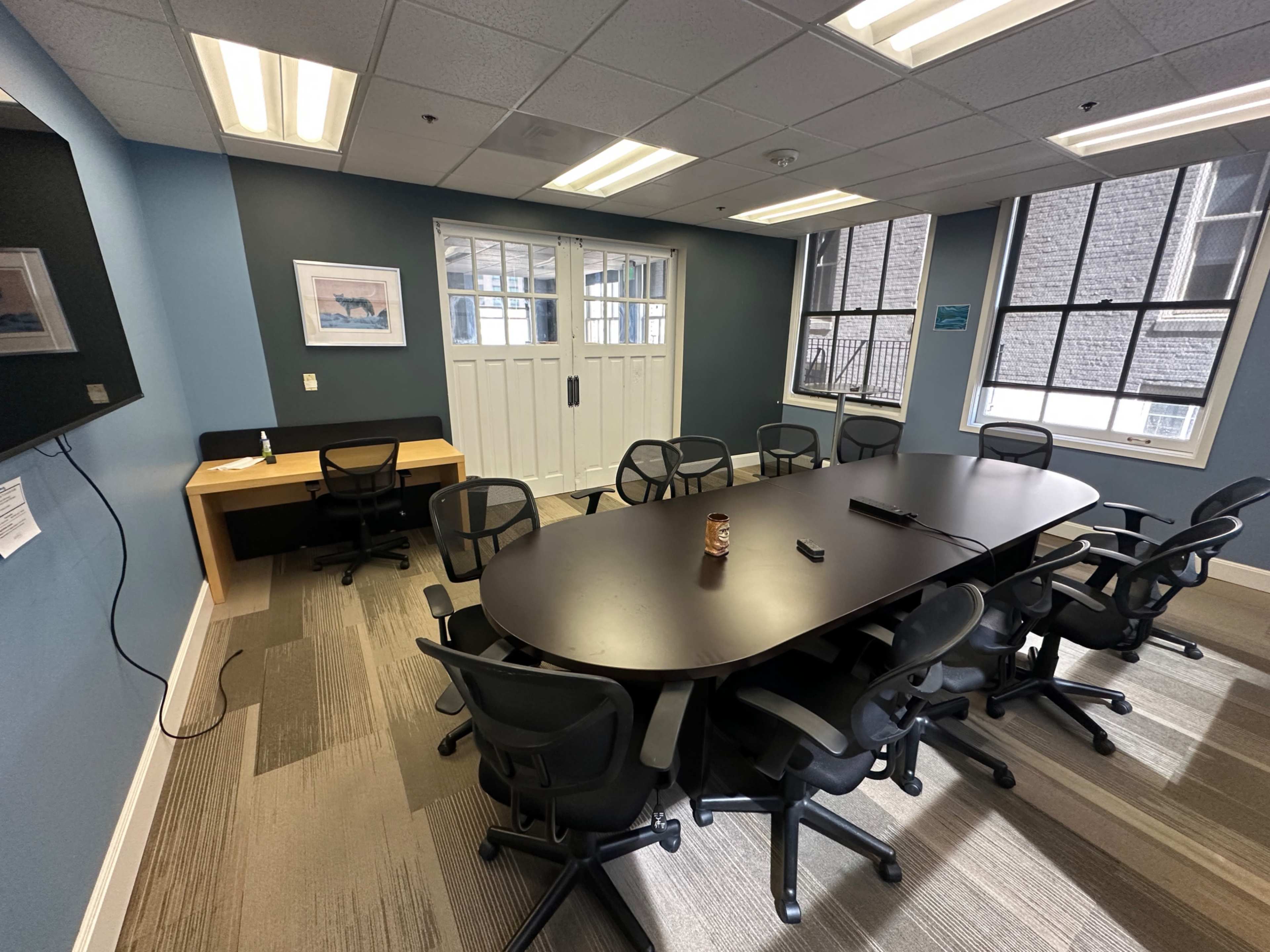 The image shows a large conference room with a black oval table surrounded by eight office chairs, a small side desk, and a wall-mounted television.