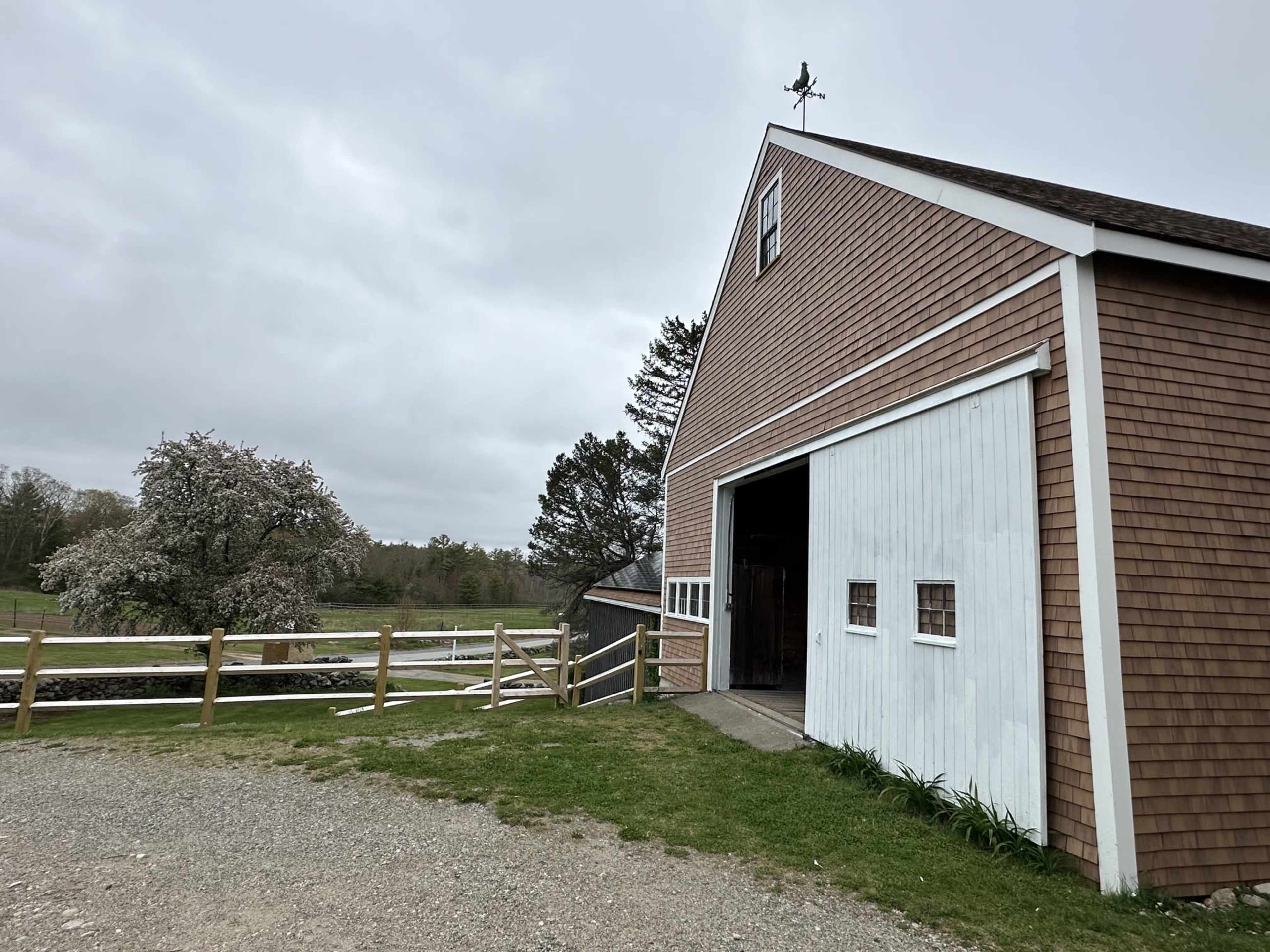 A large wooden barn with a white door is shown next to a gravel path and a grassy area, with cloudy skies in the background.