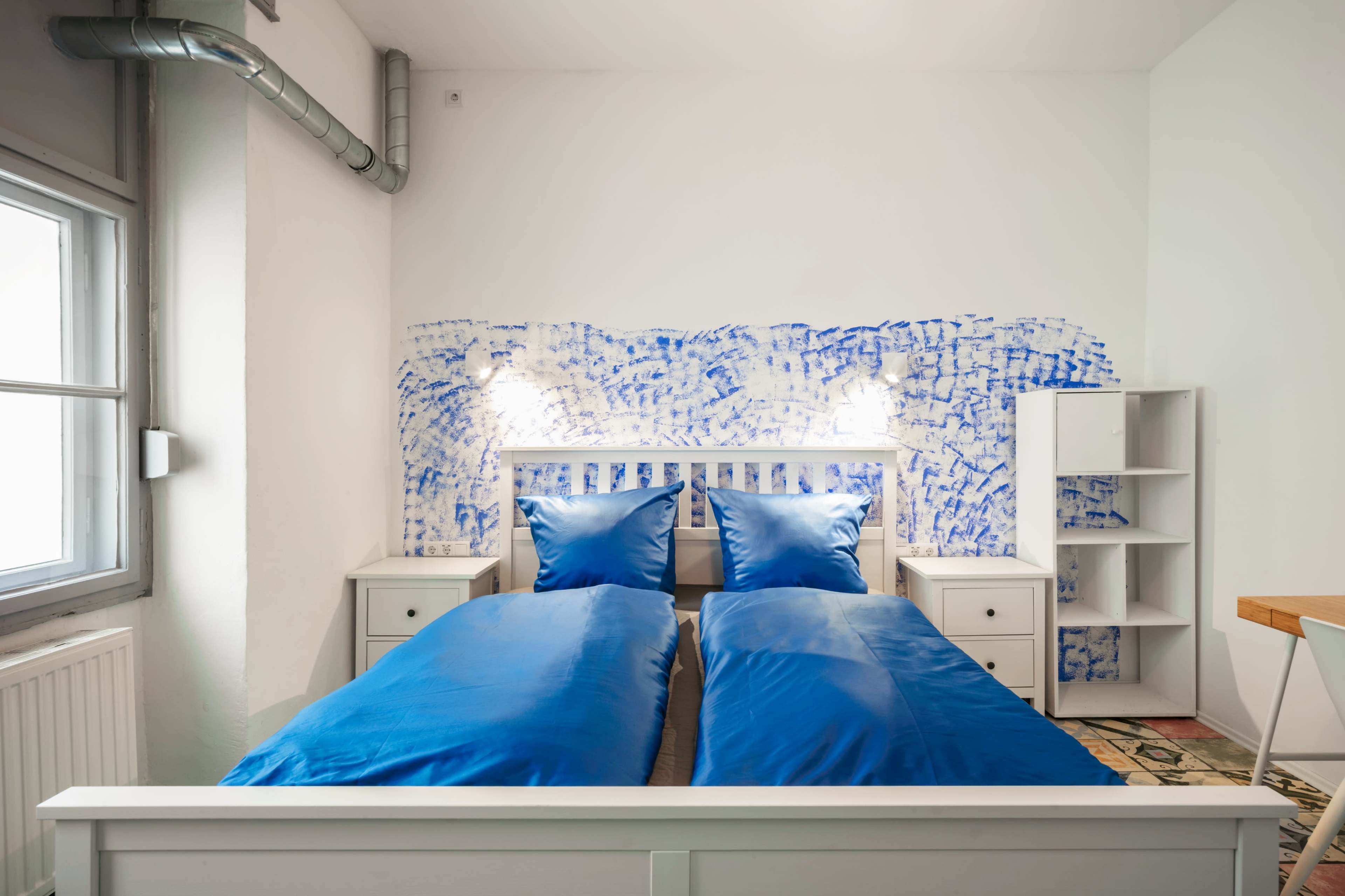 The image shows a bedroom with two twin beds featuring blue beddings, a white headboard, a shelf unit, and a wall decorated with a blue and white design.
