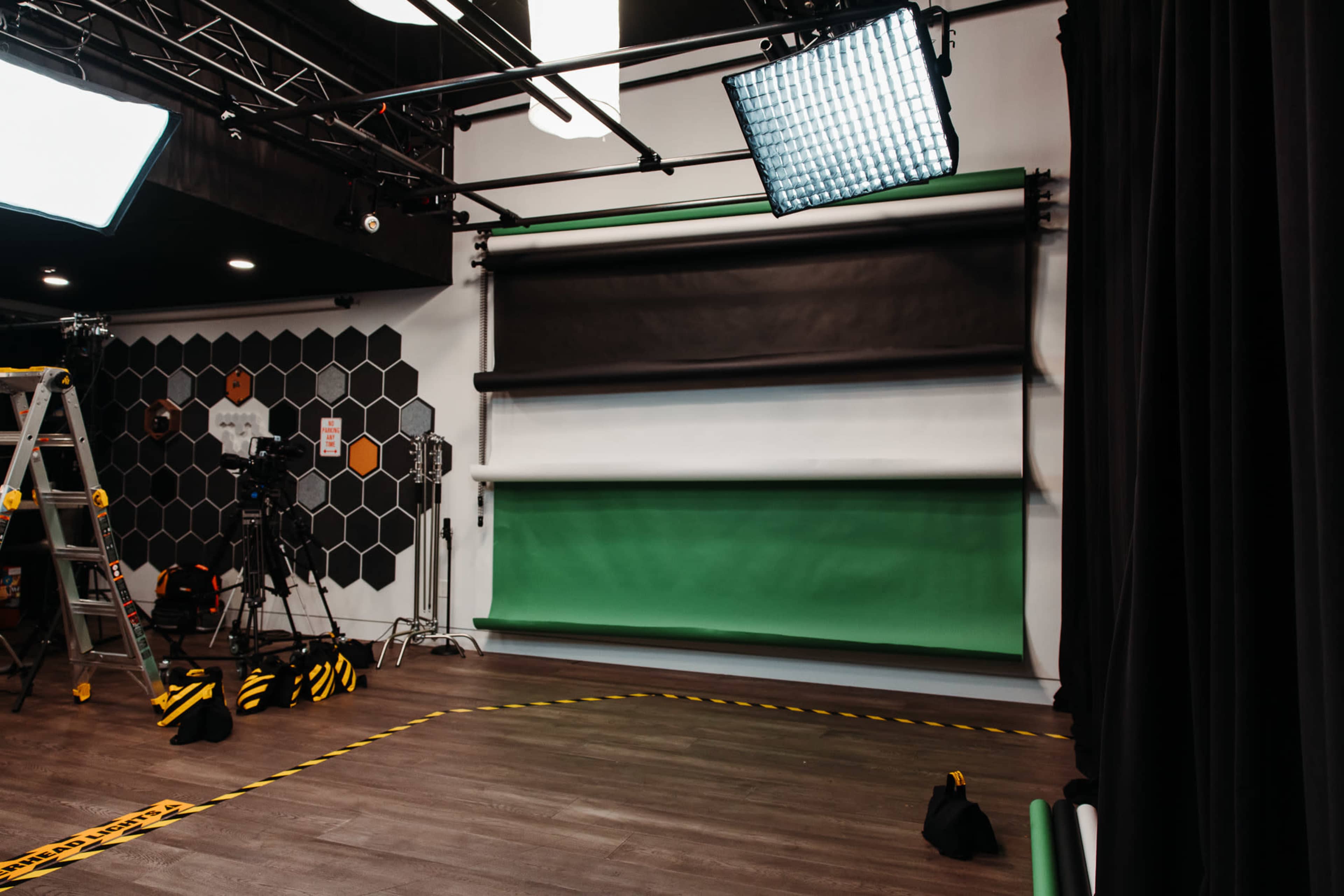 The image shows a studio setup with a green screen backdrop, studio lights, a camera on a tripod, and a ladder against a wall with a hexagonal pattern.