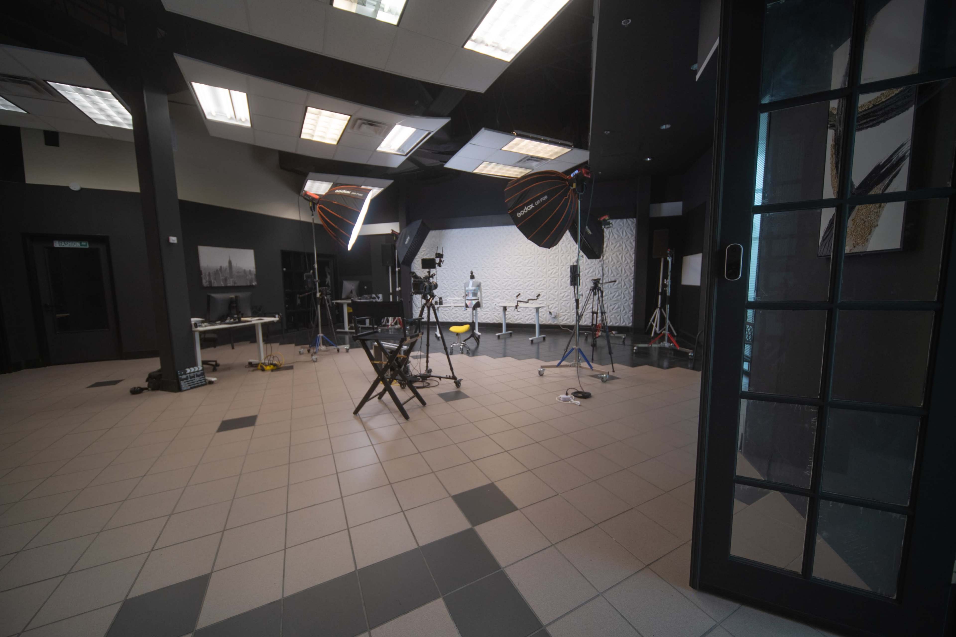 The image shows a photo studio setup with lights and cameras arranged in a spacious room featuring a tiled floor and black walls.