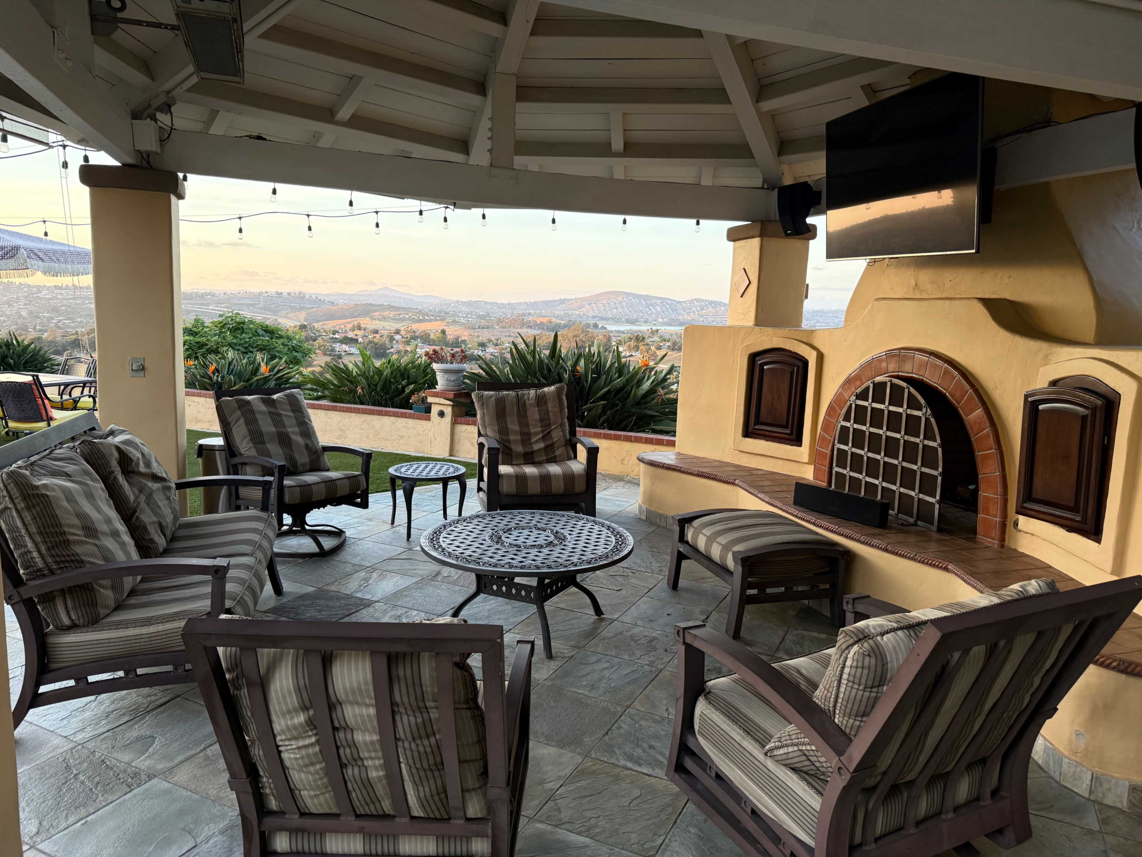 The image shows a covered outdoor seating area with several chairs and a fire pit, overlooking a distant landscape at sunset.