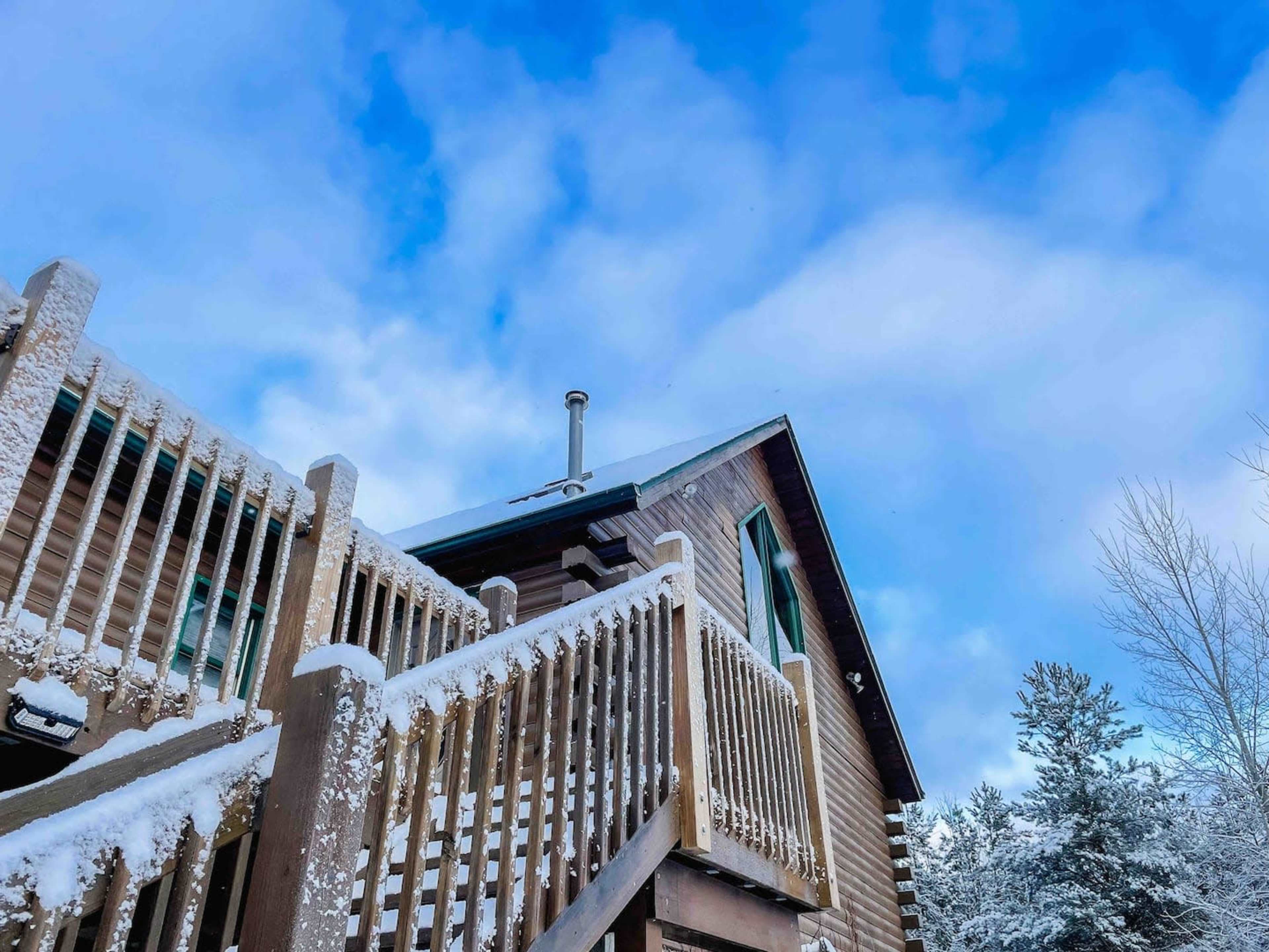 A wooden cabin with a staircase leading up to it is surrounded by snow and trees under a cloudy blue sky.