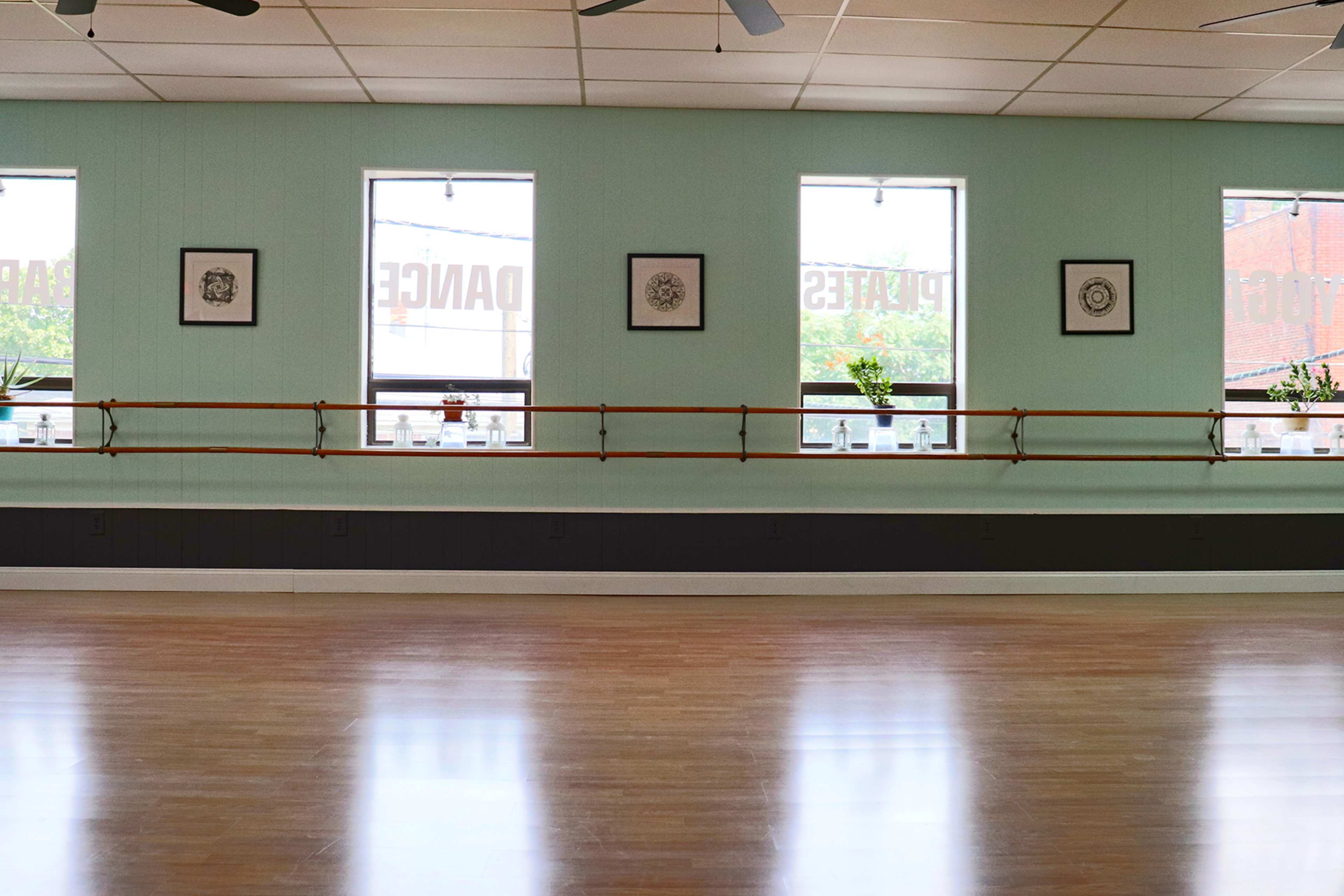 A dance studio with wooden floors, large windows featuring framed artwork, and a mirrored wall with a barre.