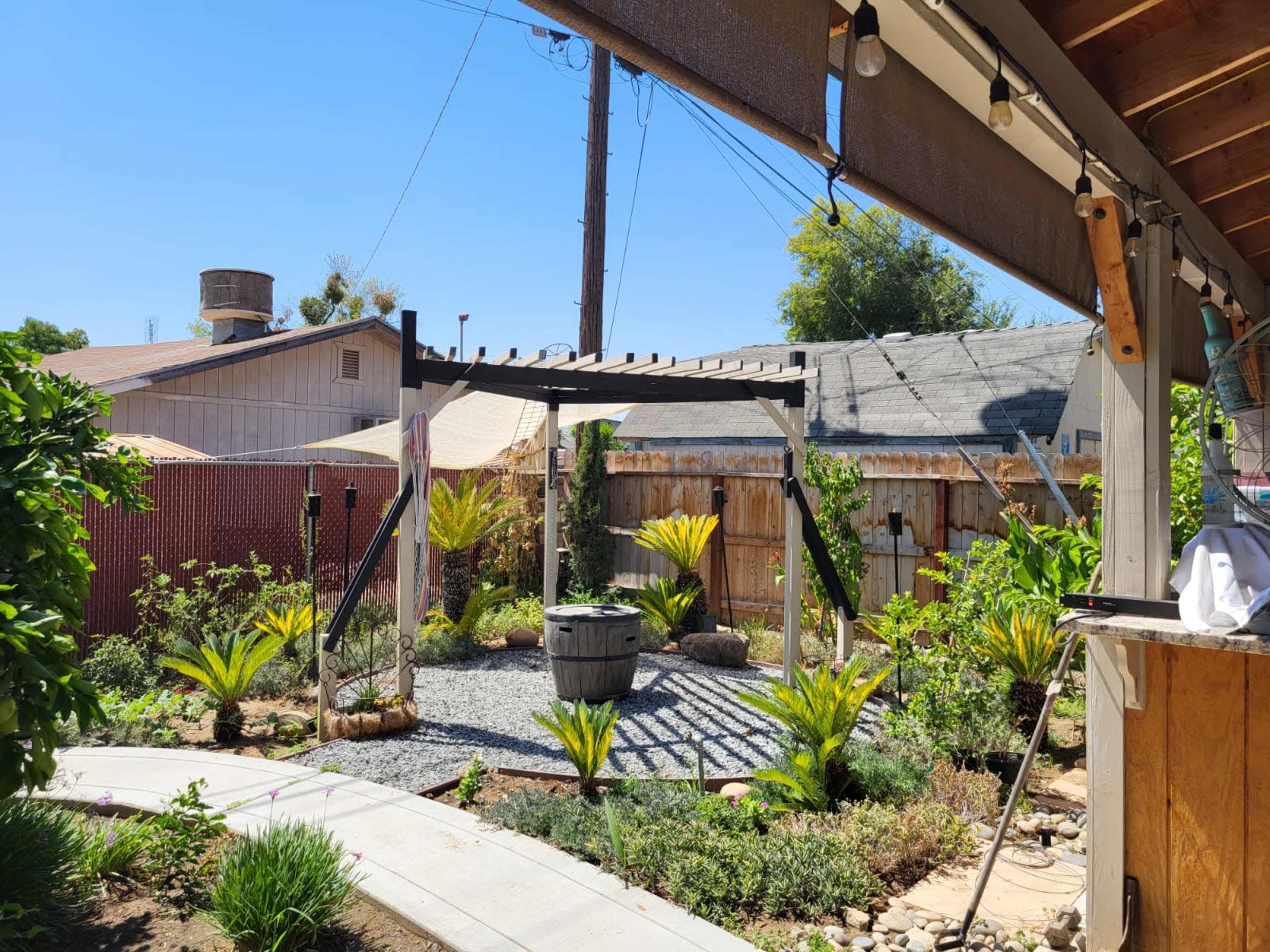 A landscaped backyard features a gravel pathway, a pergola with seating, and various plants surrounded by fences and nearby structures.