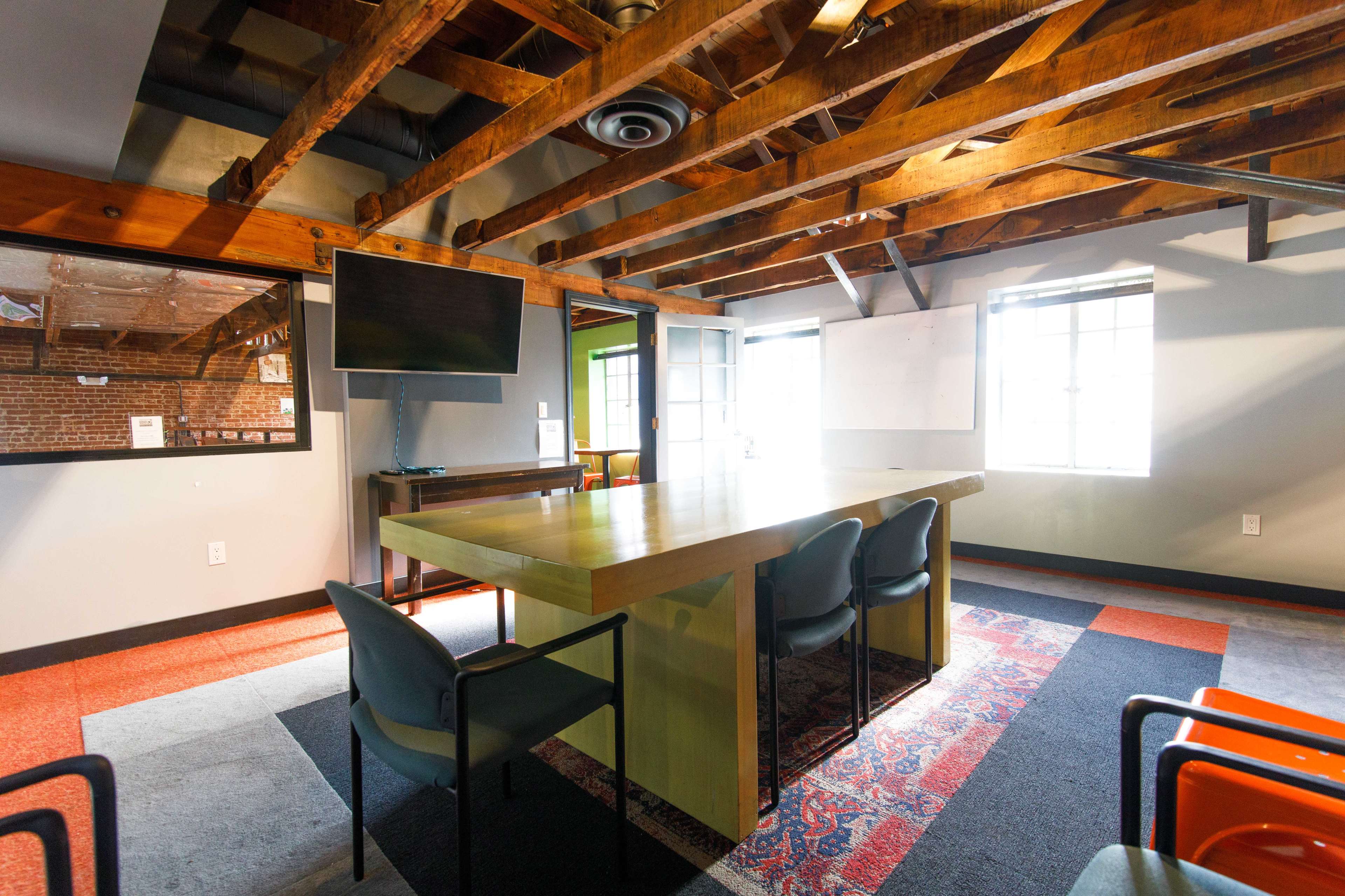 A modern meeting room featuring a long table, chairs, a television mounted on the wall, and exposed wooden beams in the ceiling.
