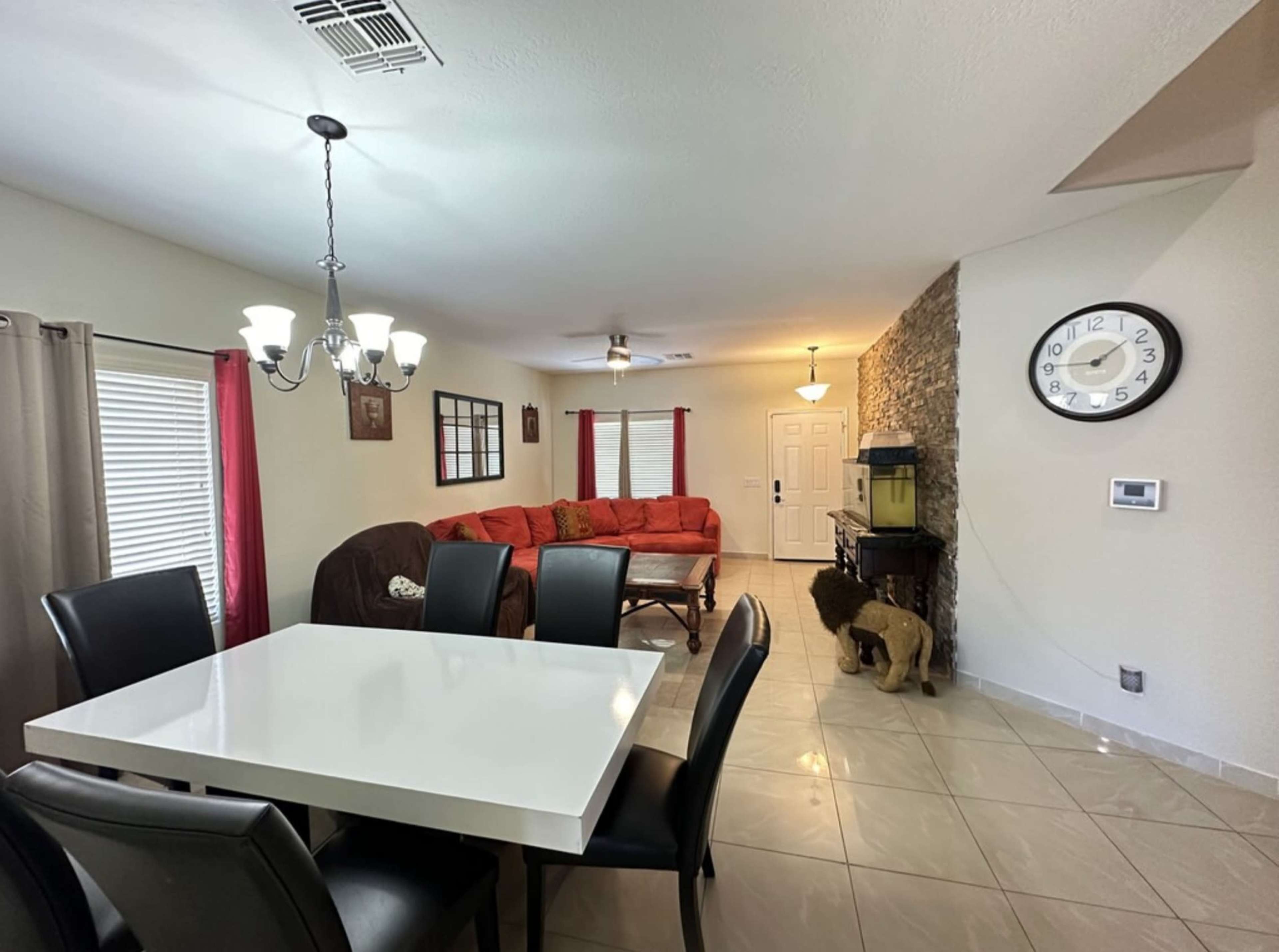 The image shows a dining area with a white table and black chairs, connected to a living room featuring a red couch, a stone accent wall, and a decorative lion statue.