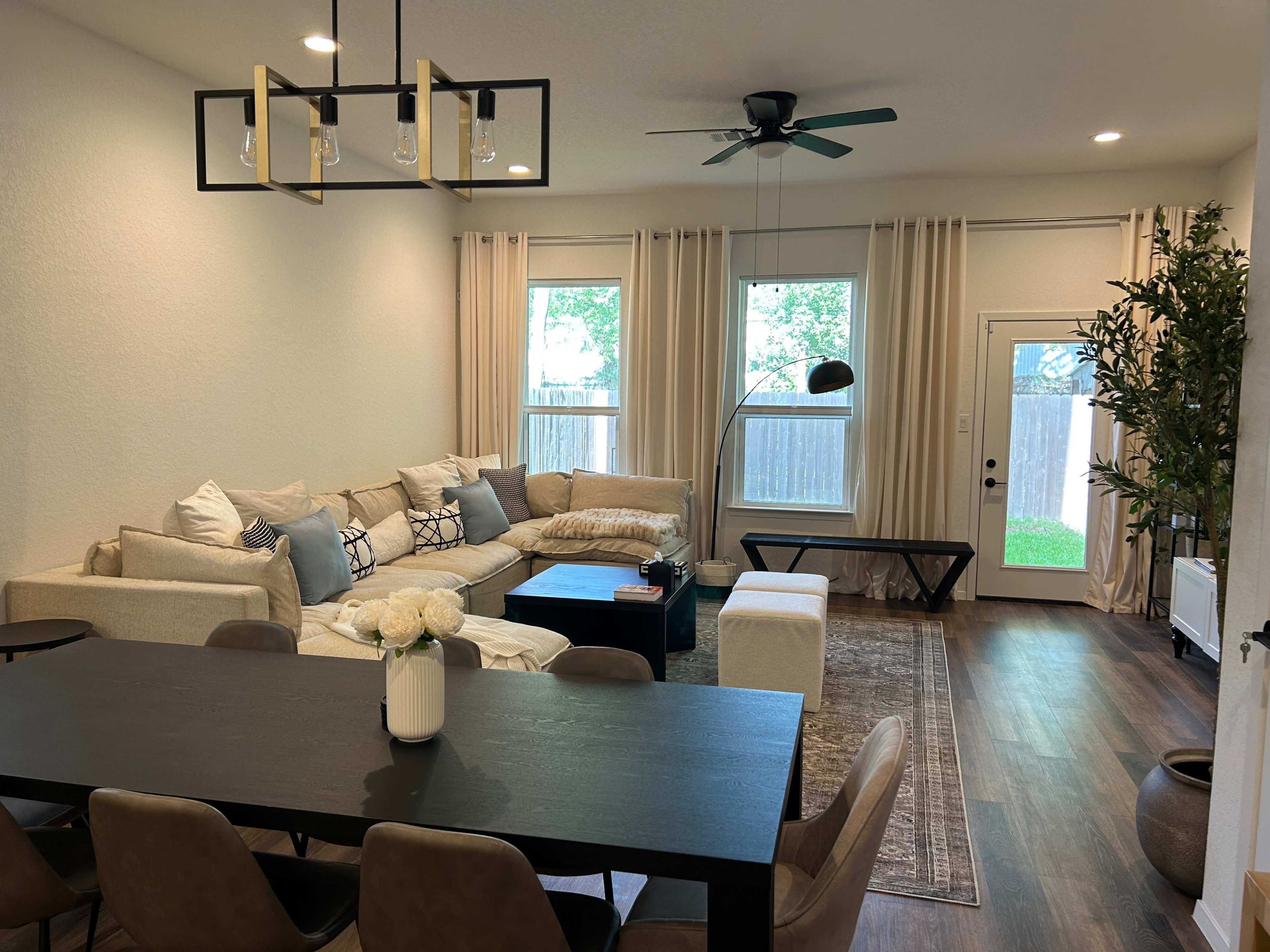 A living room with a sectional sofa, a dining table with chairs, and large windows providing natural light.