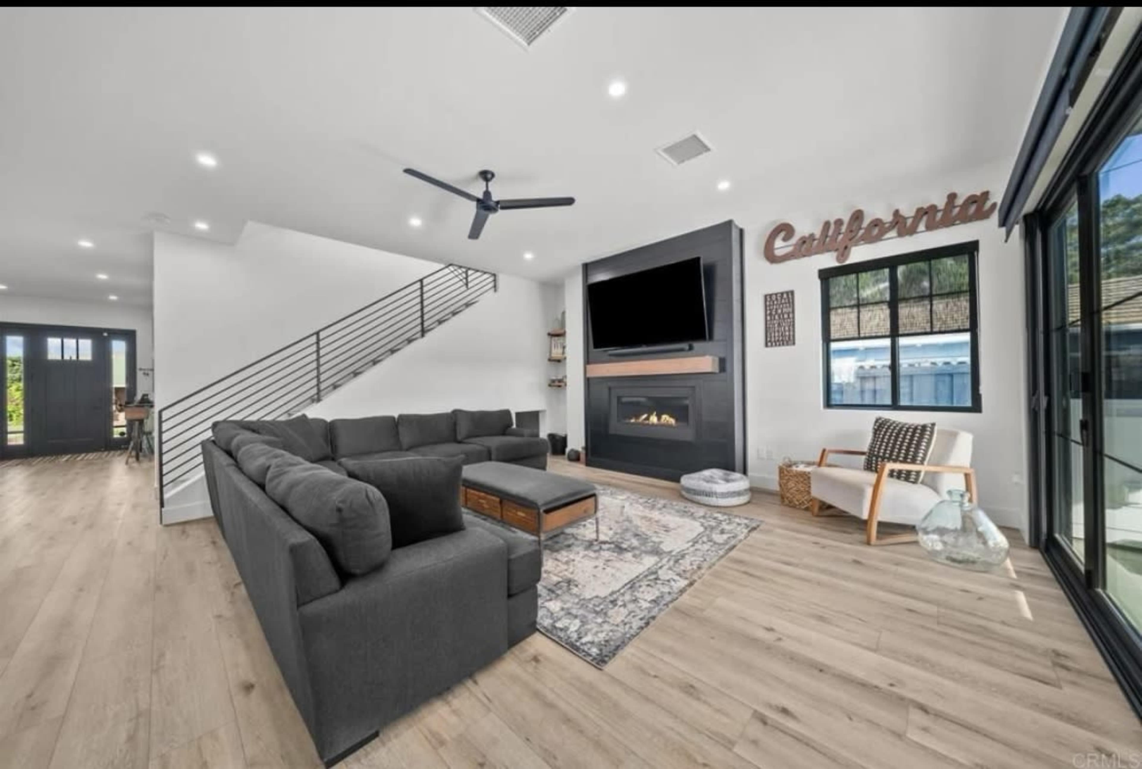 The image shows a modern living room featuring a large sectional sofa, a wall-mounted television above a fireplace, and a decorative sign that reads "Caffeinata."