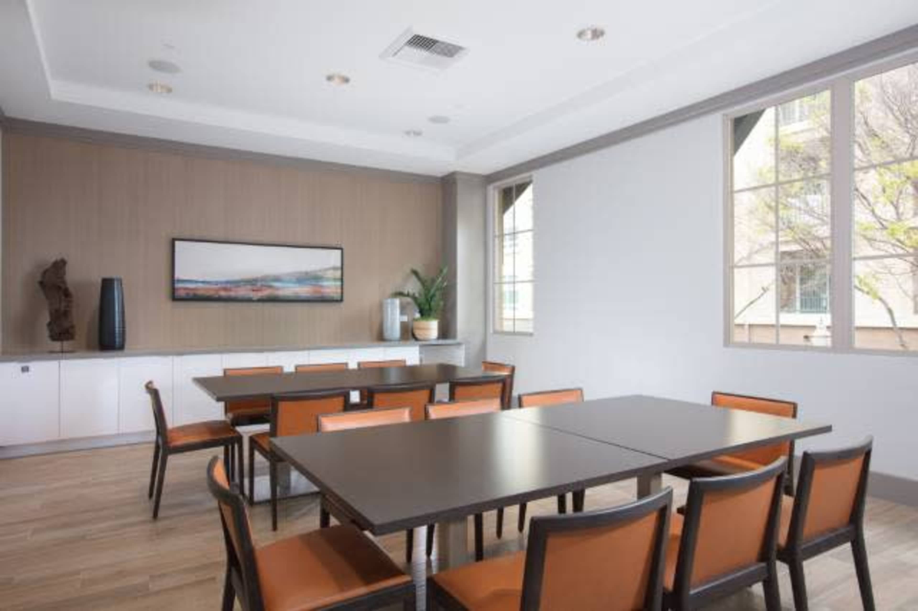 A meeting room features a large table surrounded by chairs, with a wall-mounted artwork and a plant near a window.