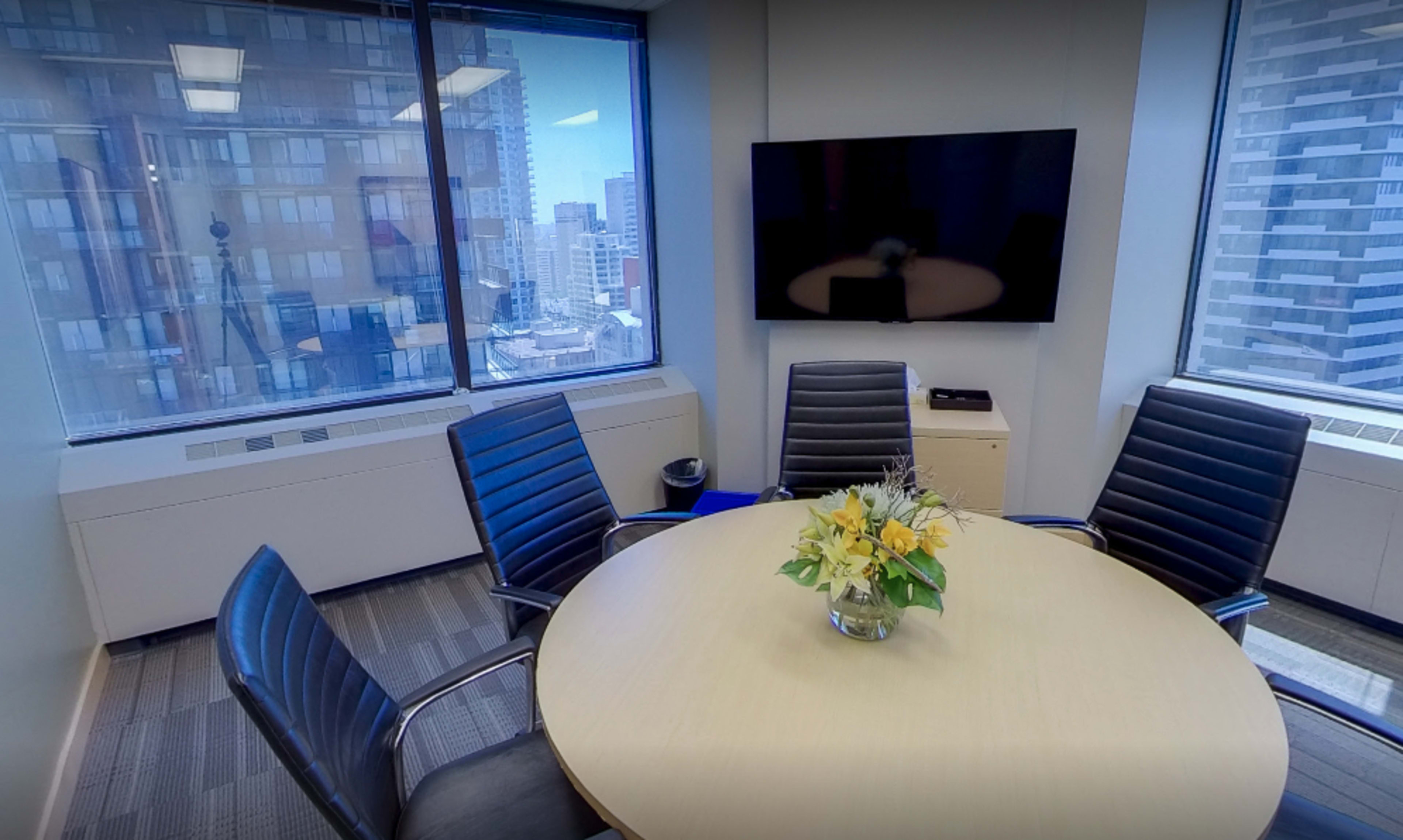 A modern conference room with a round table, five black chairs, a television on the wall, and a bouquet of flowers in the center.