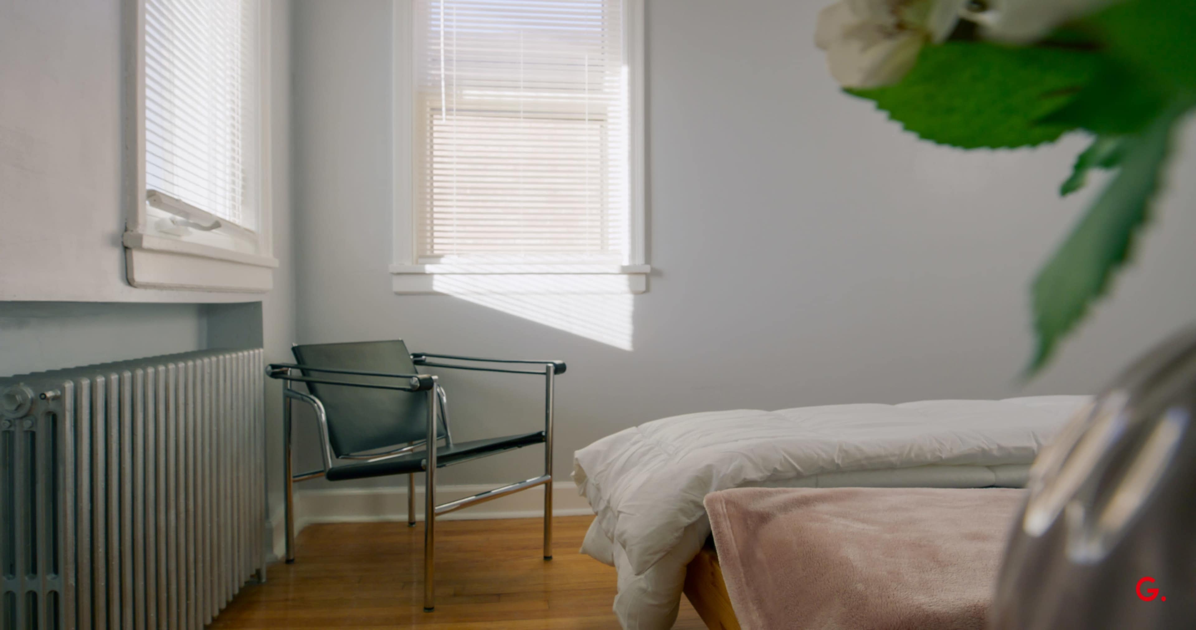 The image shows a minimalist room with a bed, a chair, and a radiator, illuminated by natural light from a window.