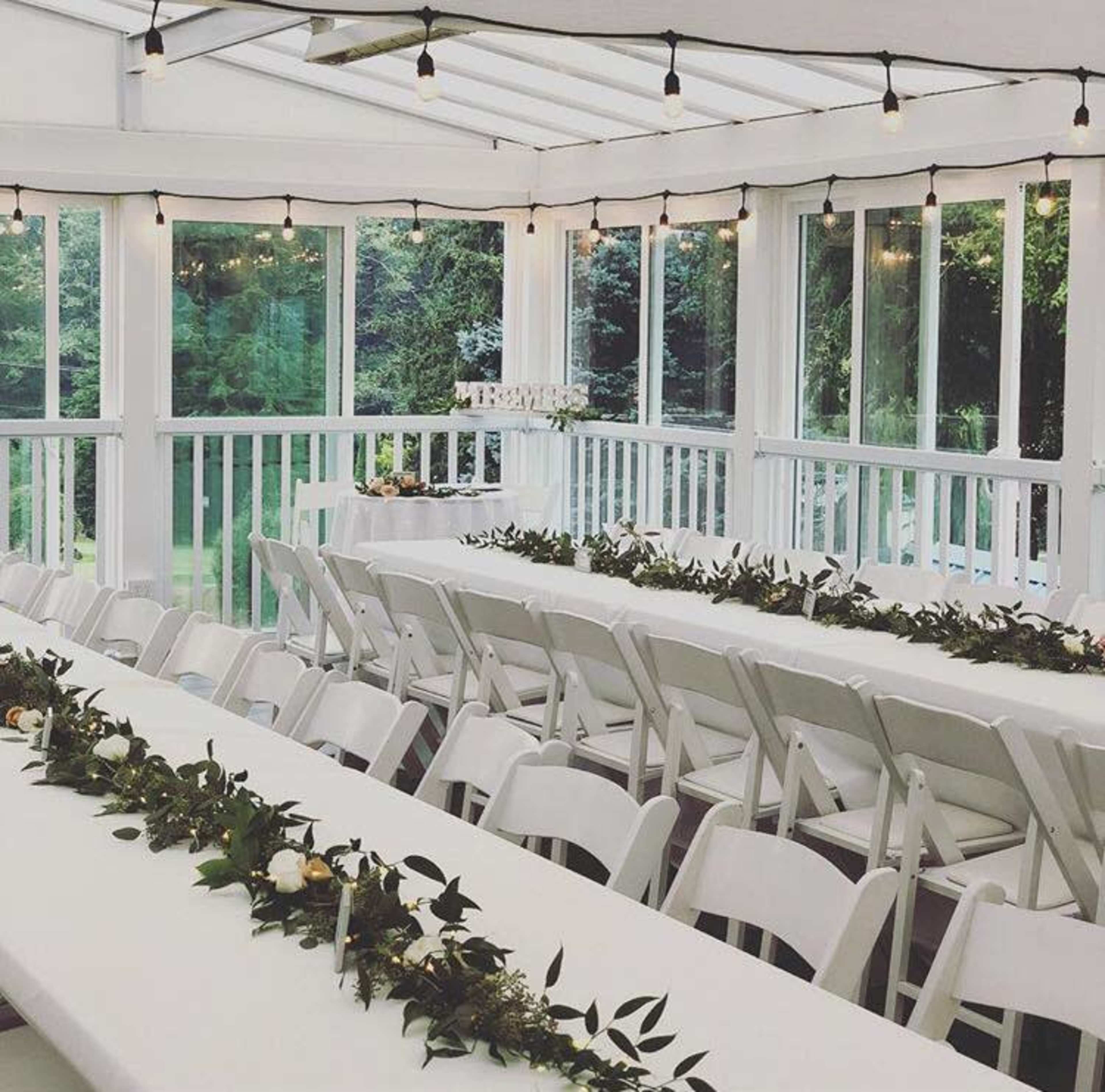 A bright room with white tables set for an event, featuring greenery centerpieces and string lights overhead.