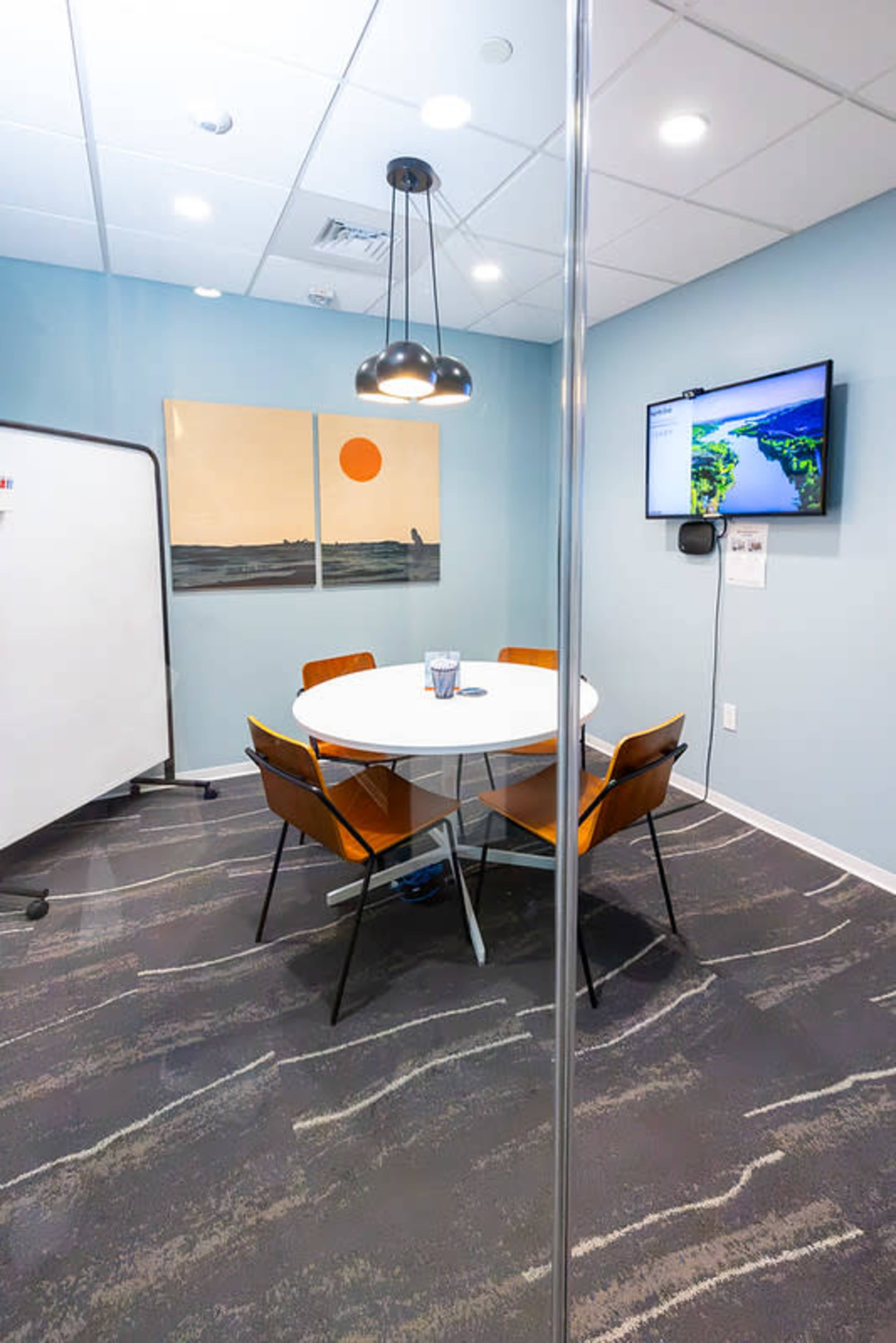 The image shows a modern conference room with a round table, four chairs, a wall-mounted TV displaying a map, and artwork featuring a sun setting over water.