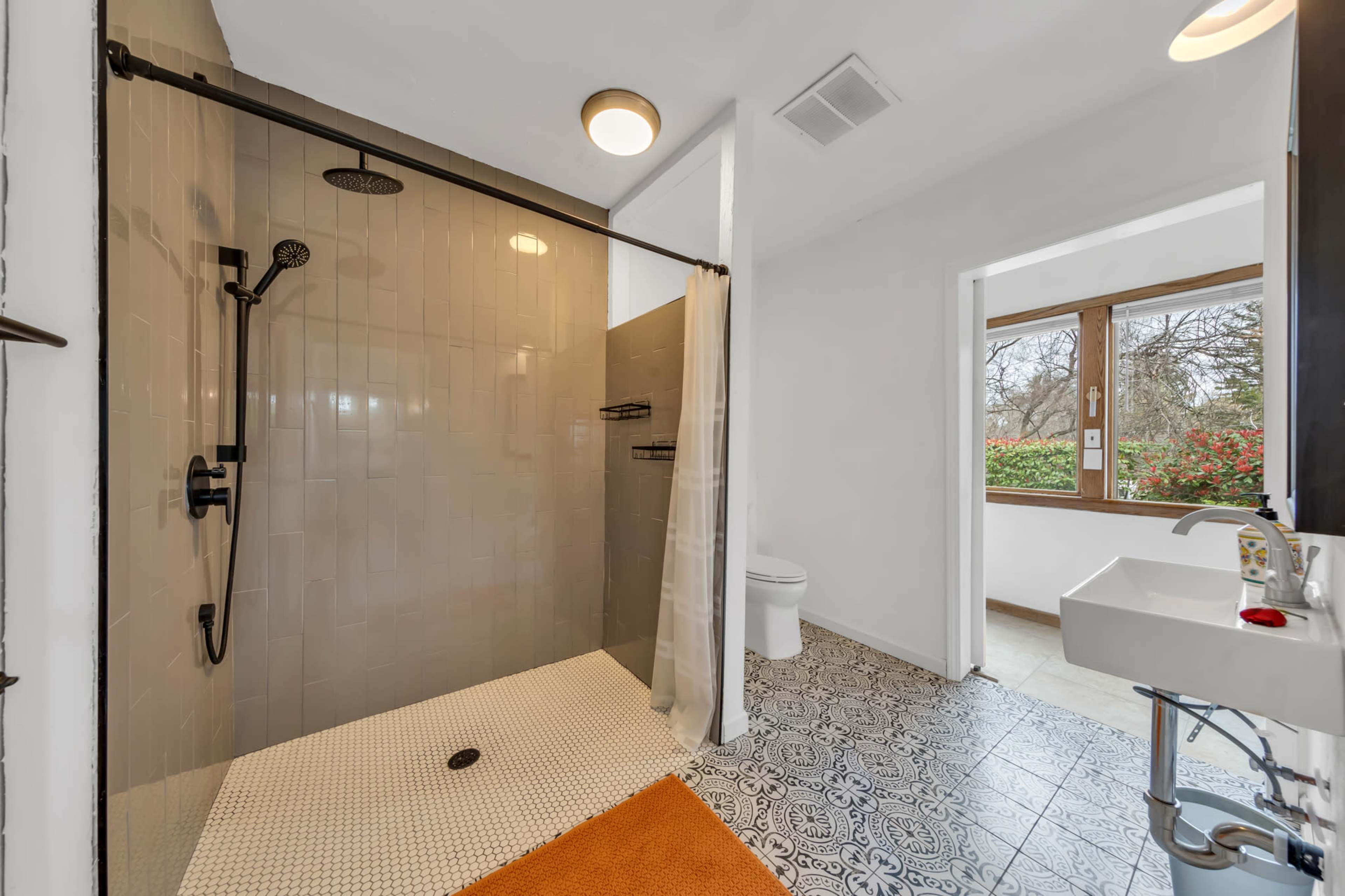 The image shows a modern bathroom featuring a walk-in shower with a glass enclosure, a toilet in the corner, and a sink beside a window.