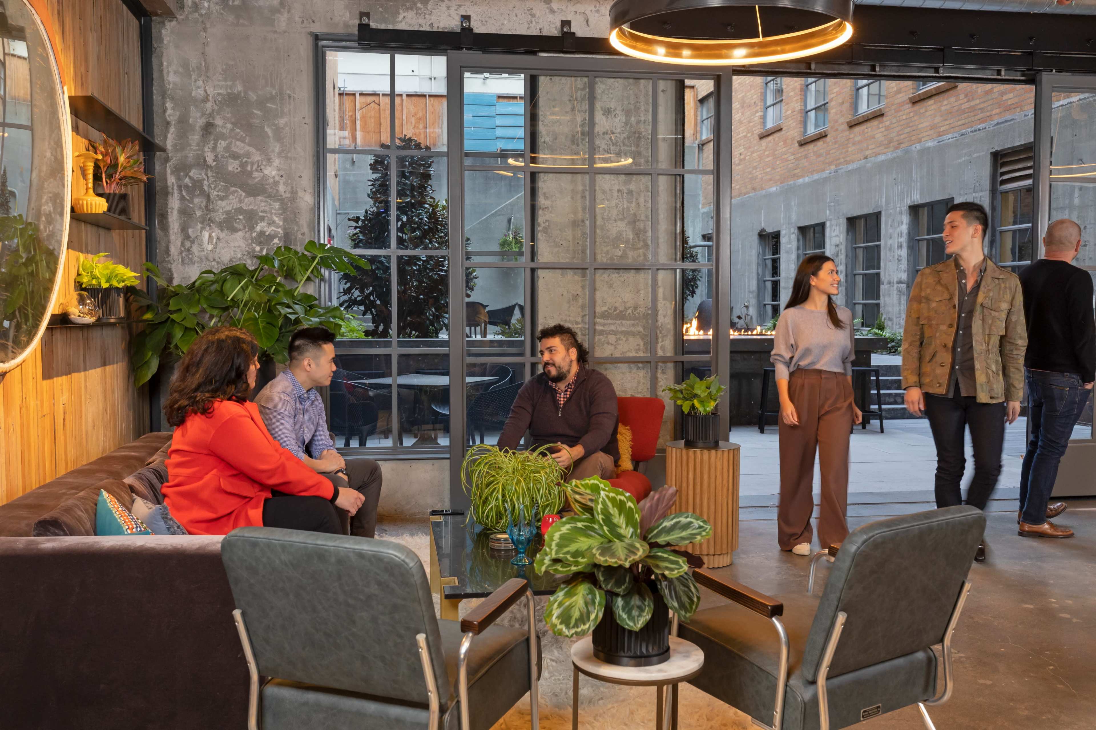 A group of people engage in conversation around a coffee table in a modern indoor space, while others walk nearby.