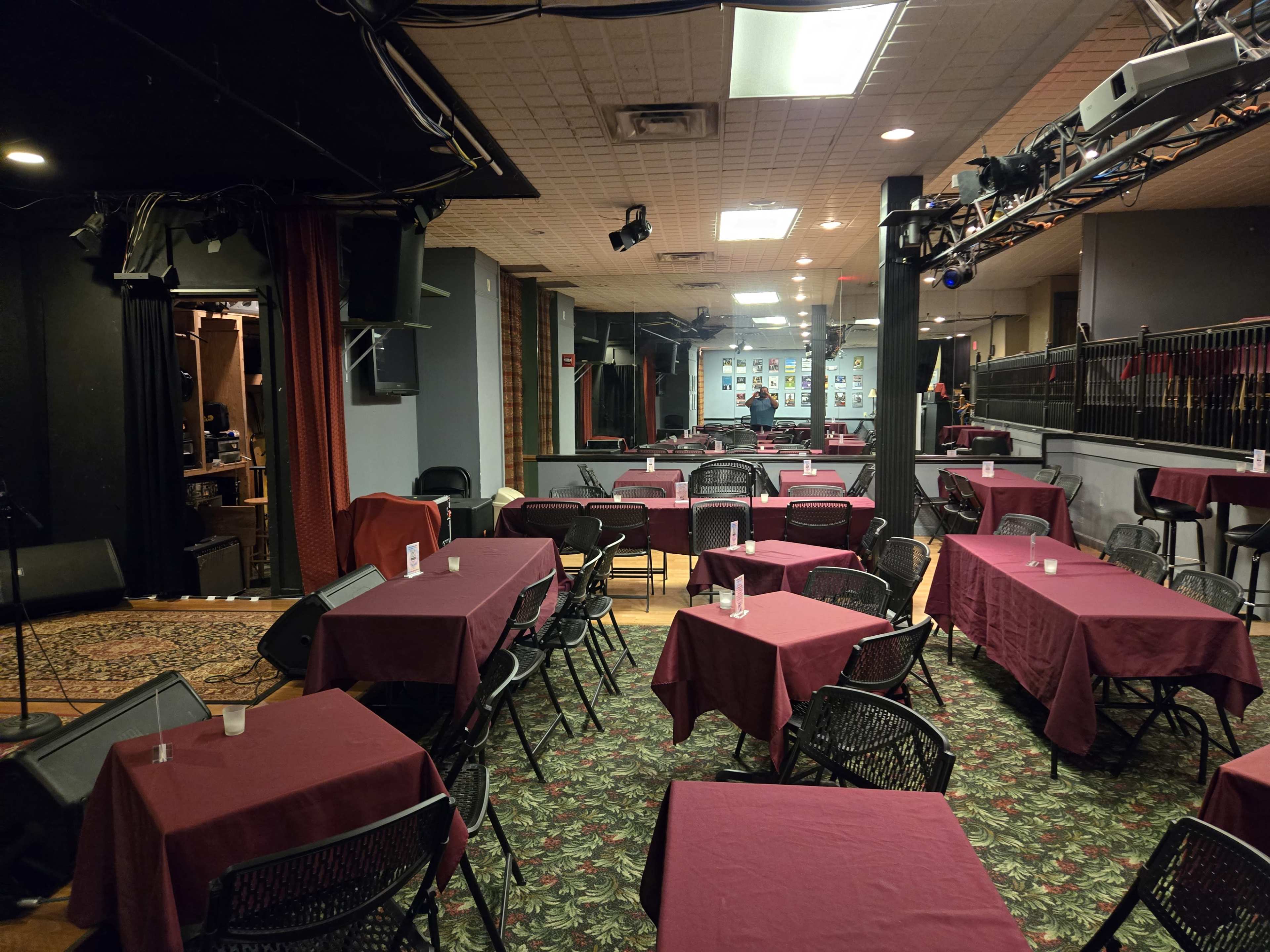 The image shows an empty performance venue with maroon tablecloths on small round tables and a stage area with sound equipment in the background.