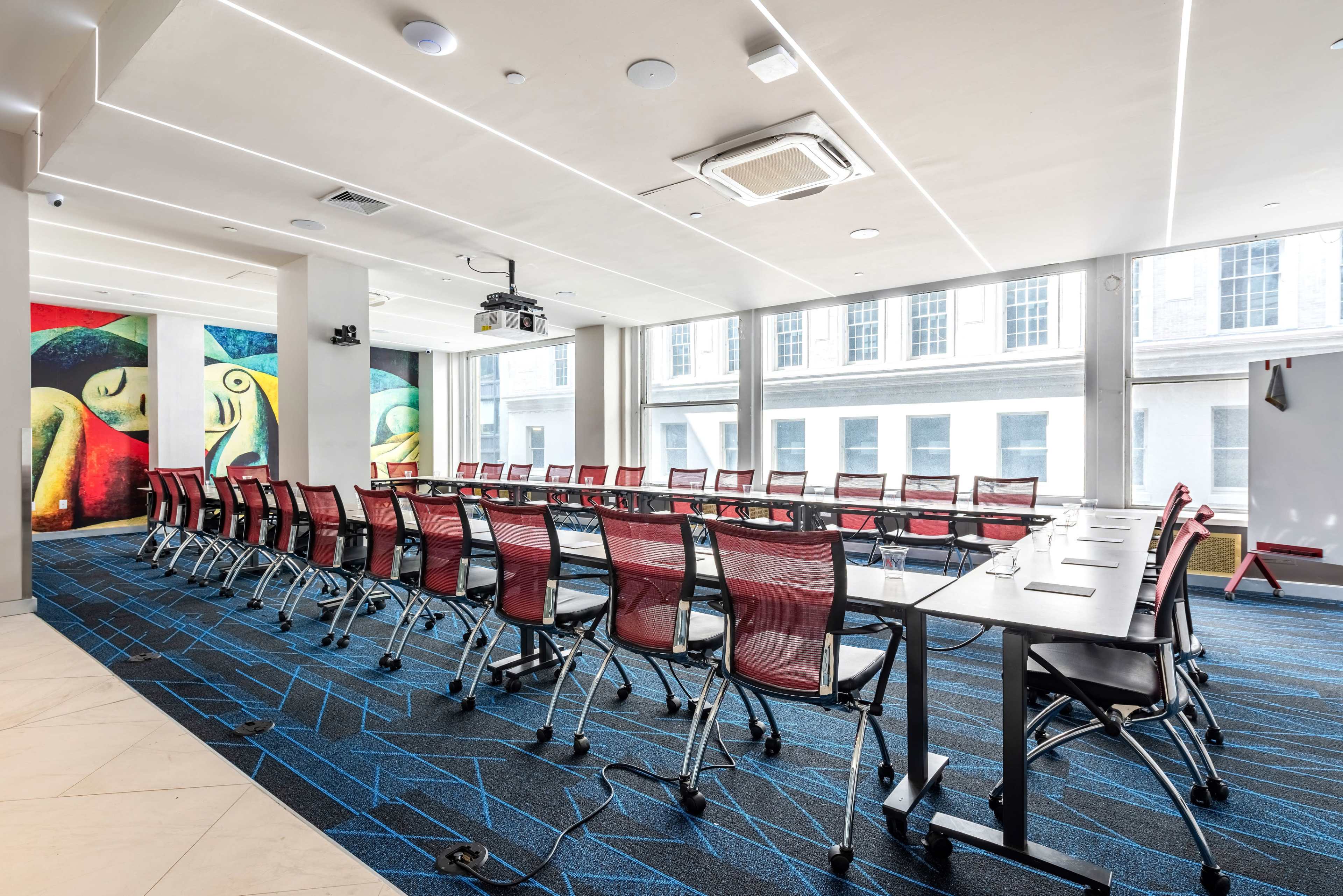 The image shows a modern conference room with a long table surrounded by red mesh chairs and colorful artwork on the walls.