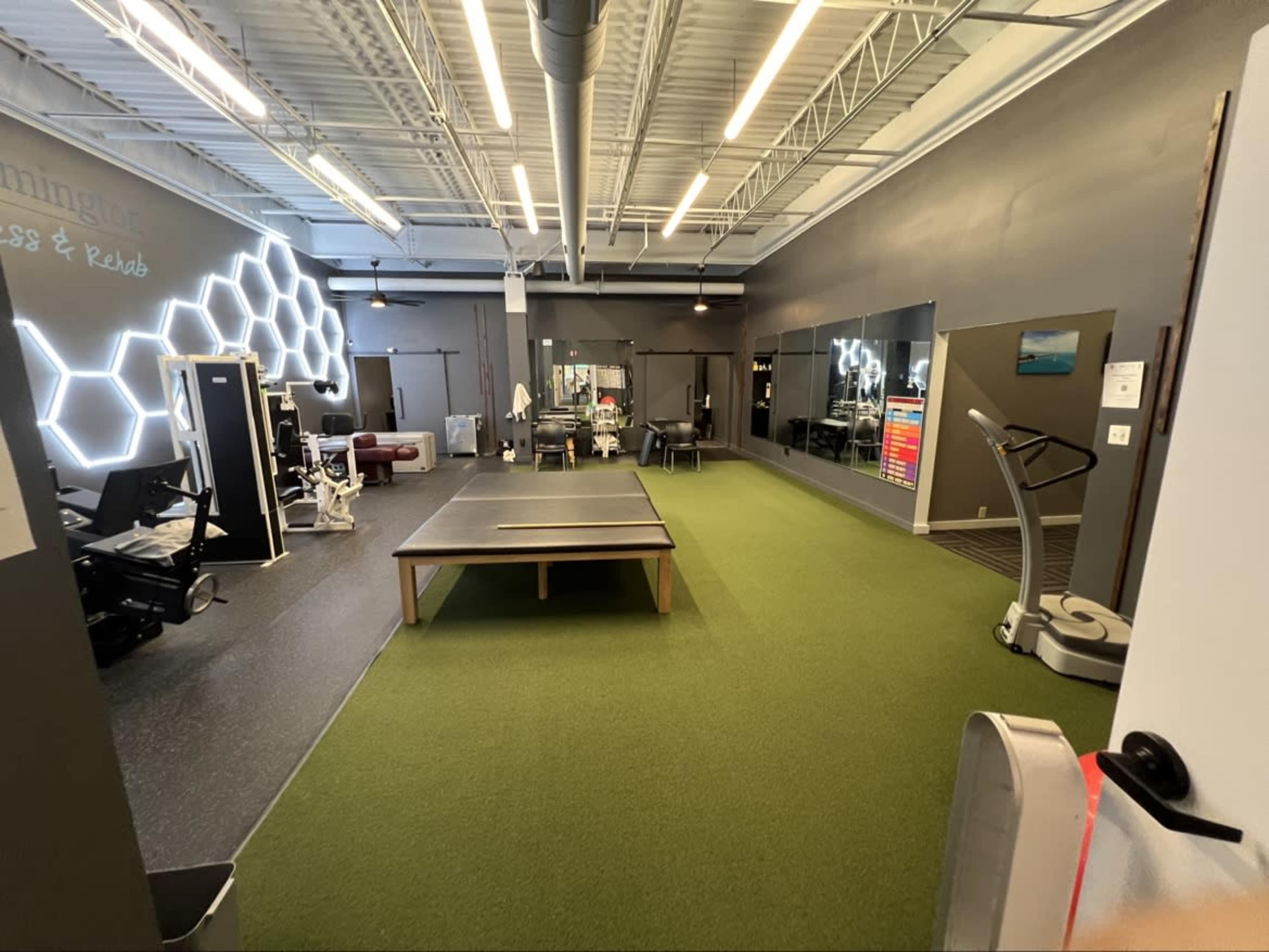 The image shows a spacious fitness and rehabilitation facility featuring green turf flooring, exercise equipment, and large mirrors on the walls.