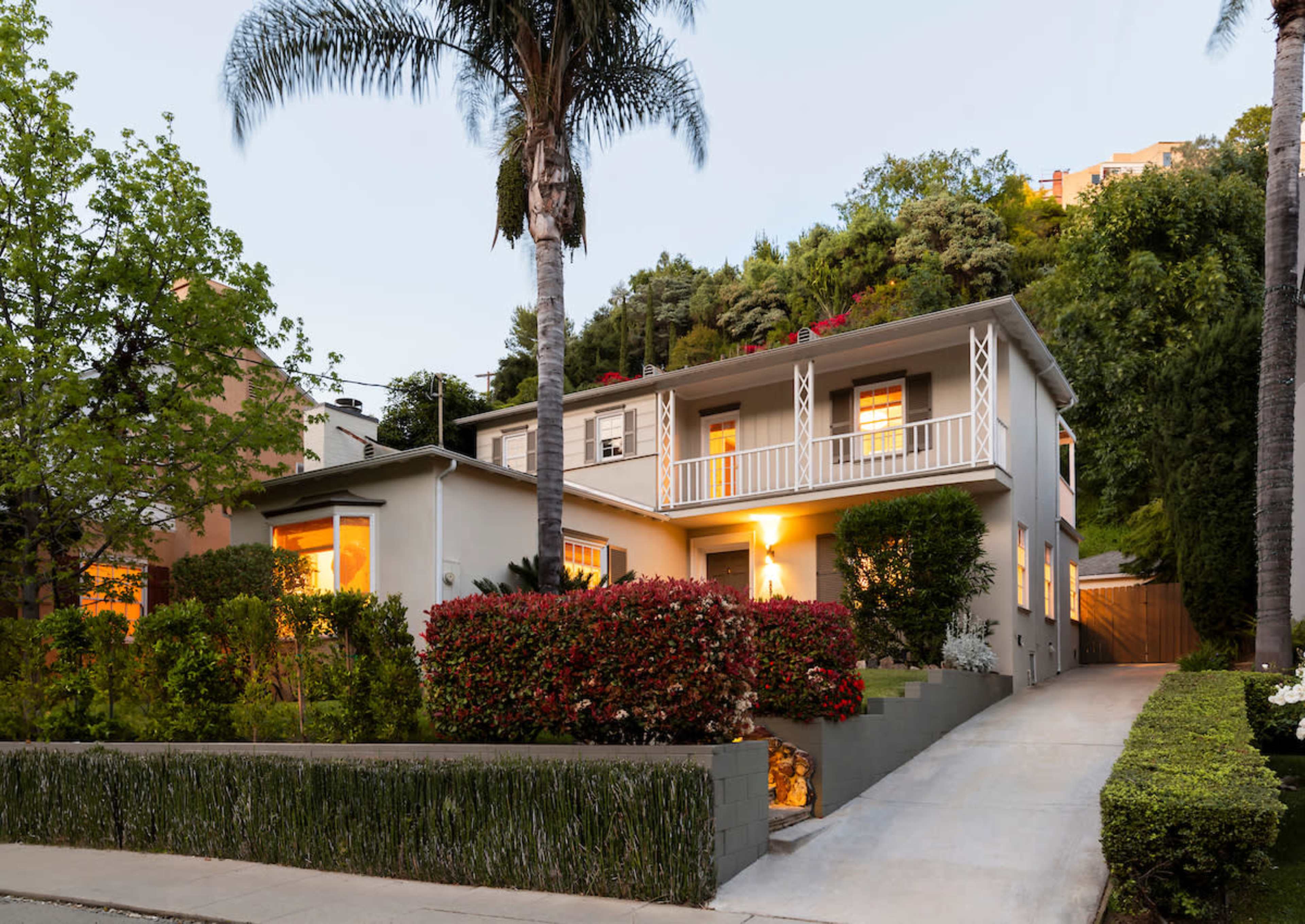 A two-story house with a landscaped yard and palm trees is illuminated in the evening.