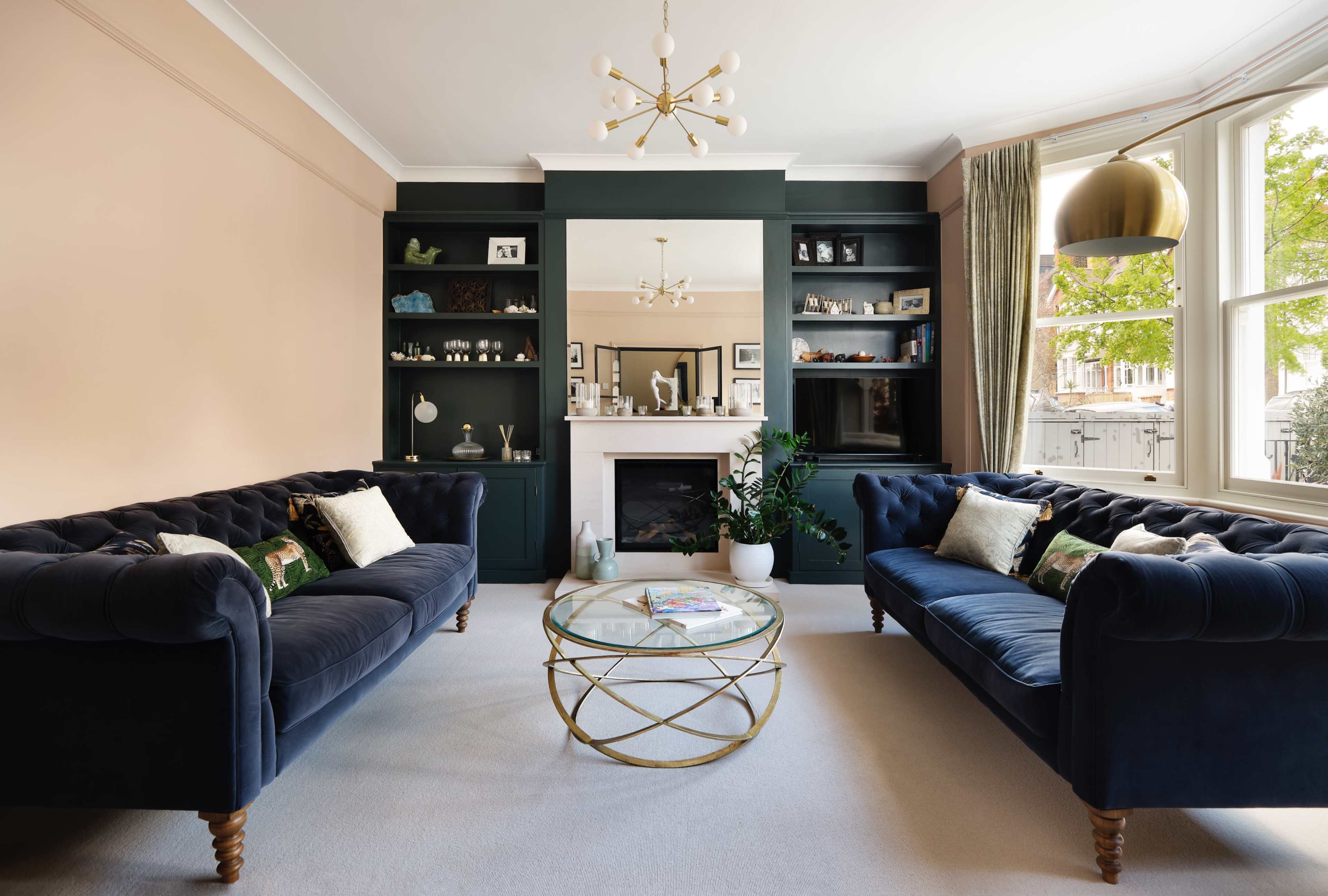 The living room features two blue velvet sofas facing a glass coffee table, with a modern chandelier and a fireplace surrounded by built-in shelves.