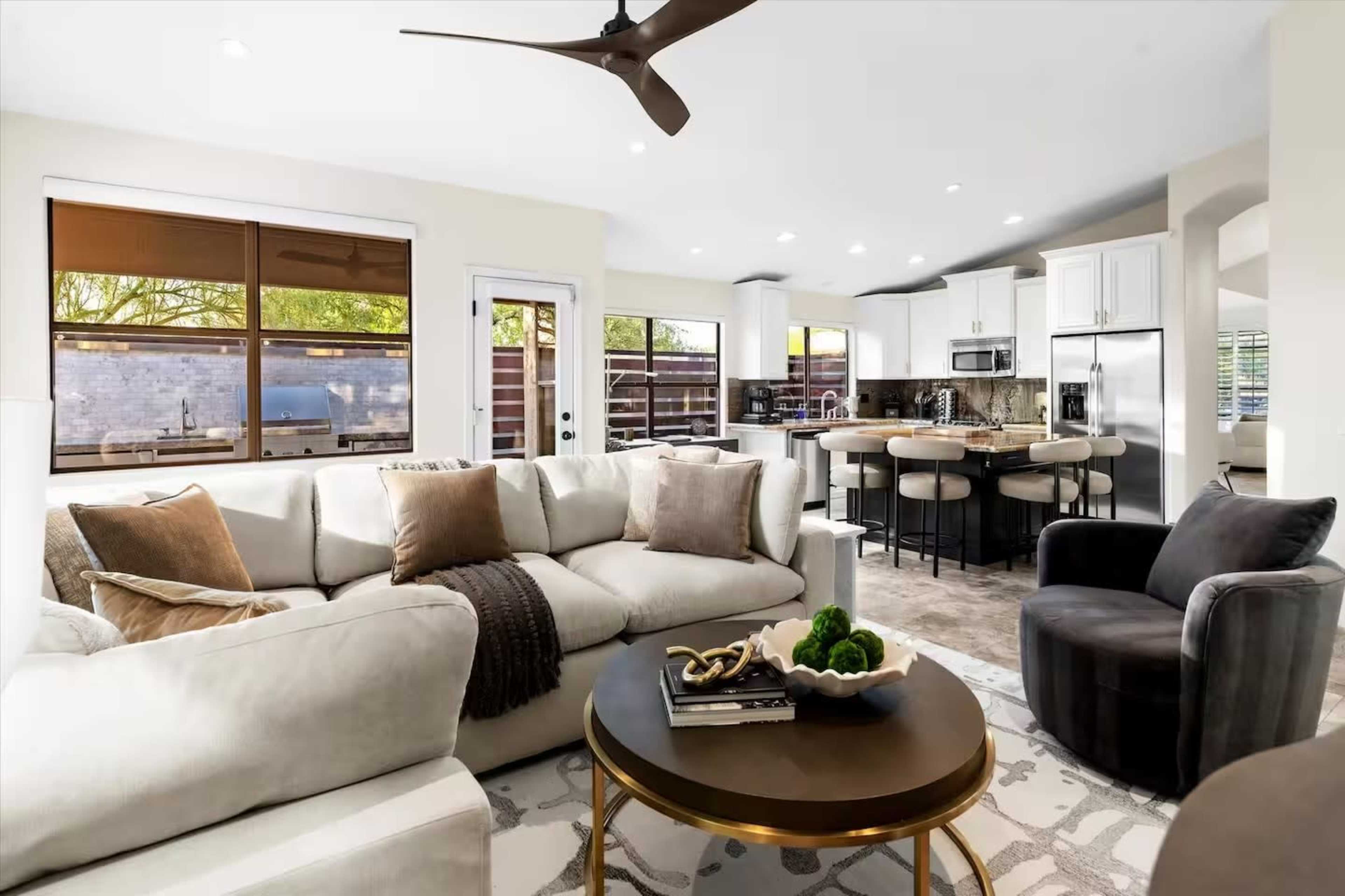 The image shows a modern living room with a large sectional sofa, a round coffee table, and an open kitchen visible in the background.