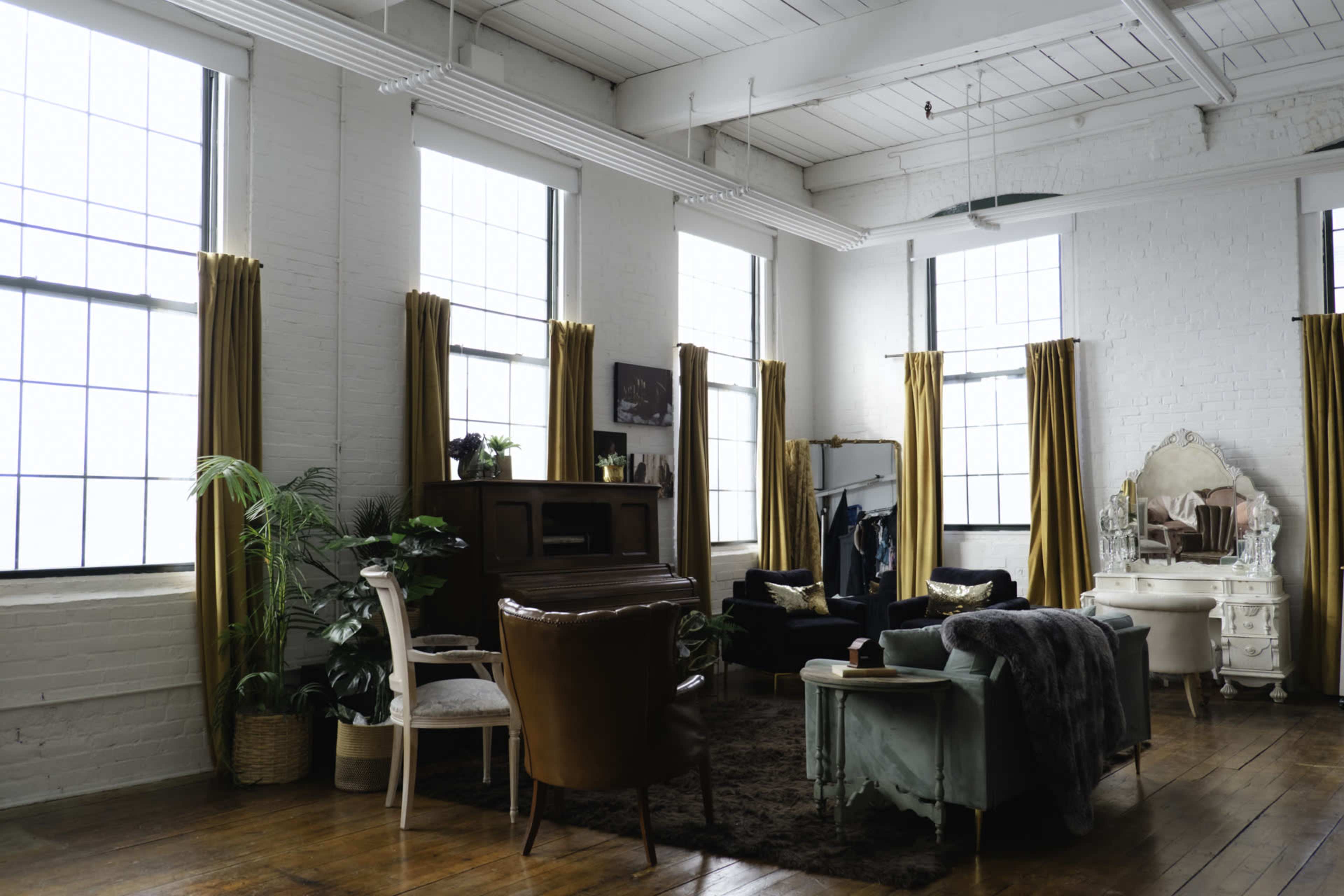 The image shows a spacious room with large windows, hardwood floors, and a mixture of seating options, including a piano and decorative furniture.